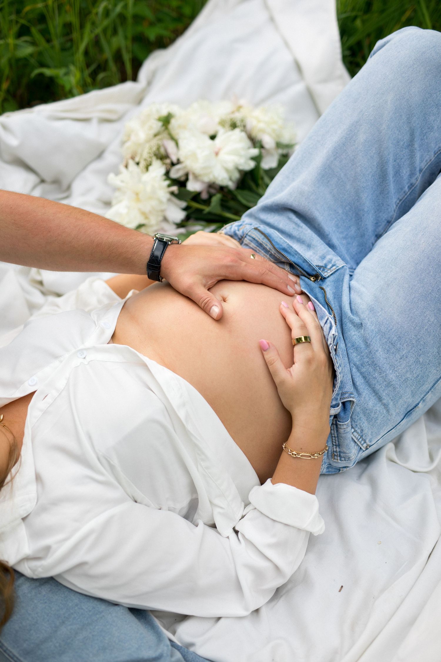 Couple tenderly touching pregnant belly outdoors on white blanket with white peonies, wearing jeans and white shirt.