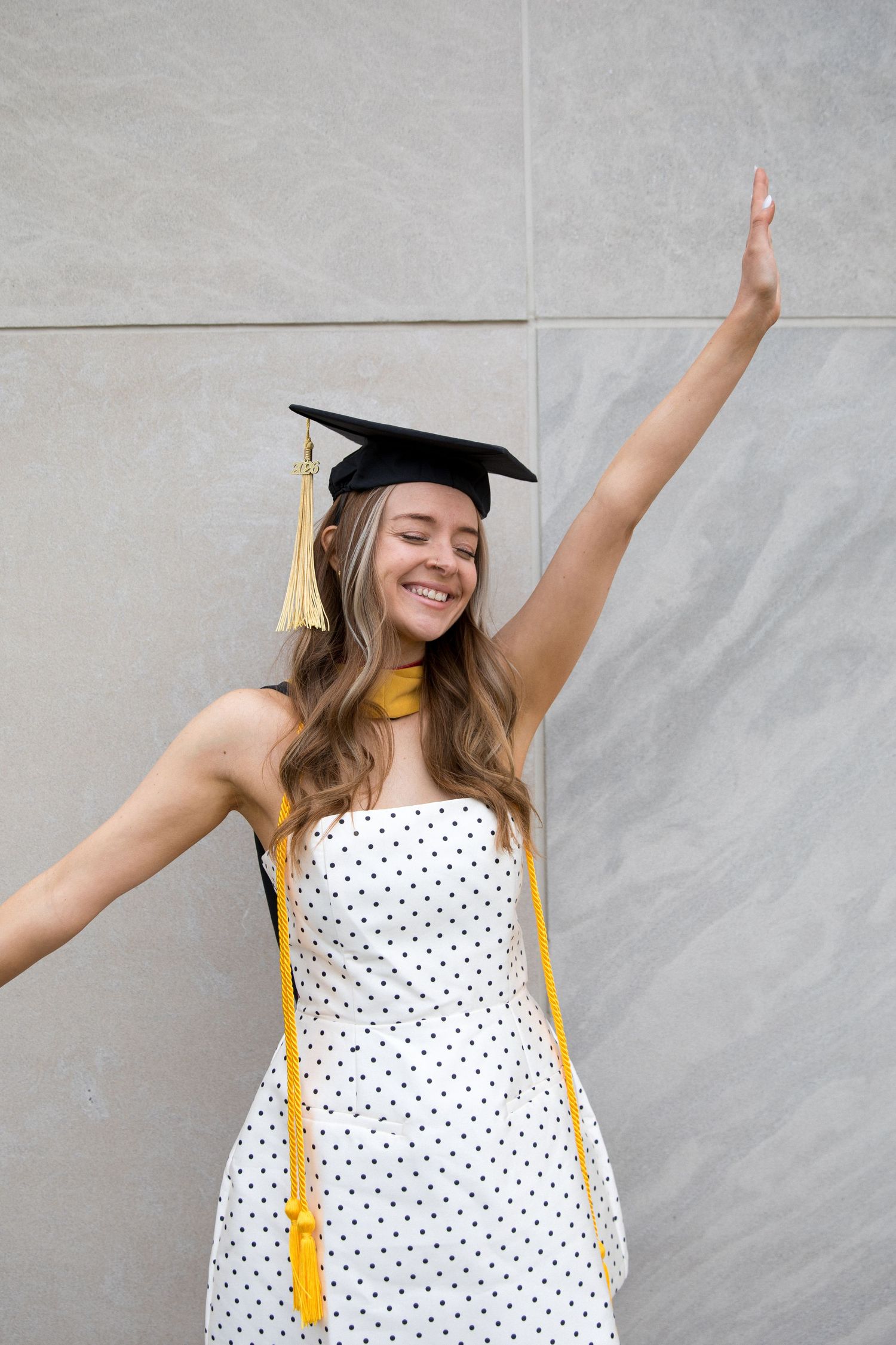 Smiling young woman in polka dot dress and graduation cap celebrating with arm raised against gray wall.