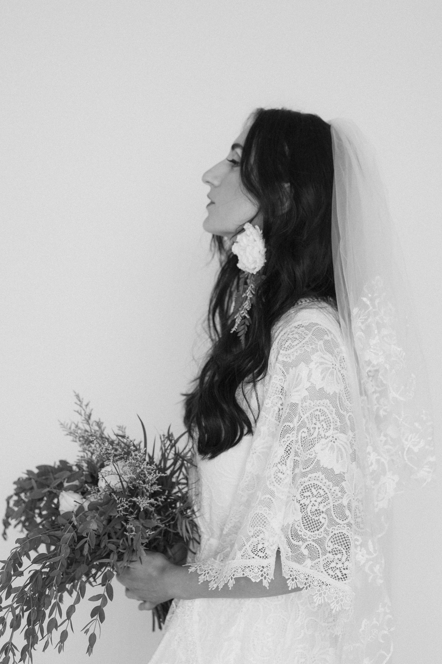 Black and white bridal portrait of a woman in lace wedding dress and veil holding a bouquet, side profile view.