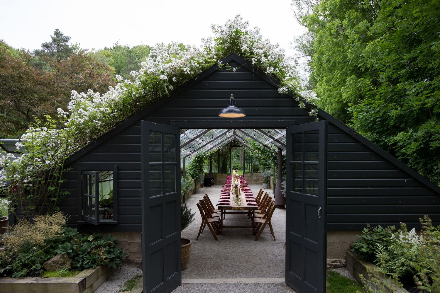 A rustic black wooden garden shed with climbing vines and a covered seating area in a lush green setting.