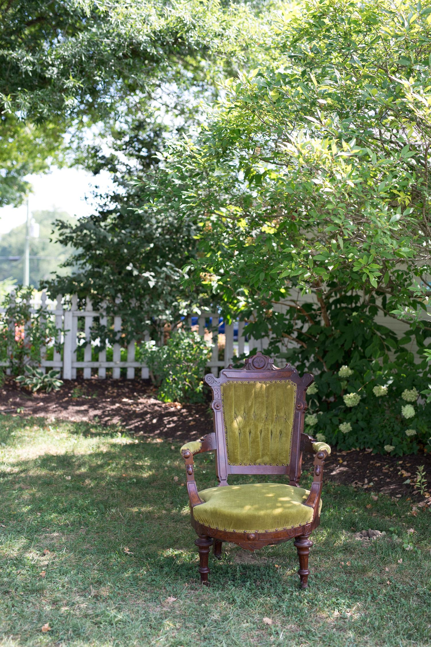 Antique green velvet armchair sitting on grass in a lush garden with white picket fence and flowering shrubs.