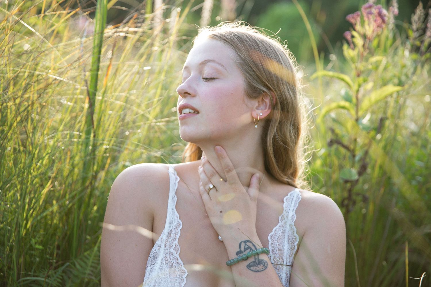 A person in a white lace dress poses thoughtfully in a sunlit field of tall grass and wildflowers.