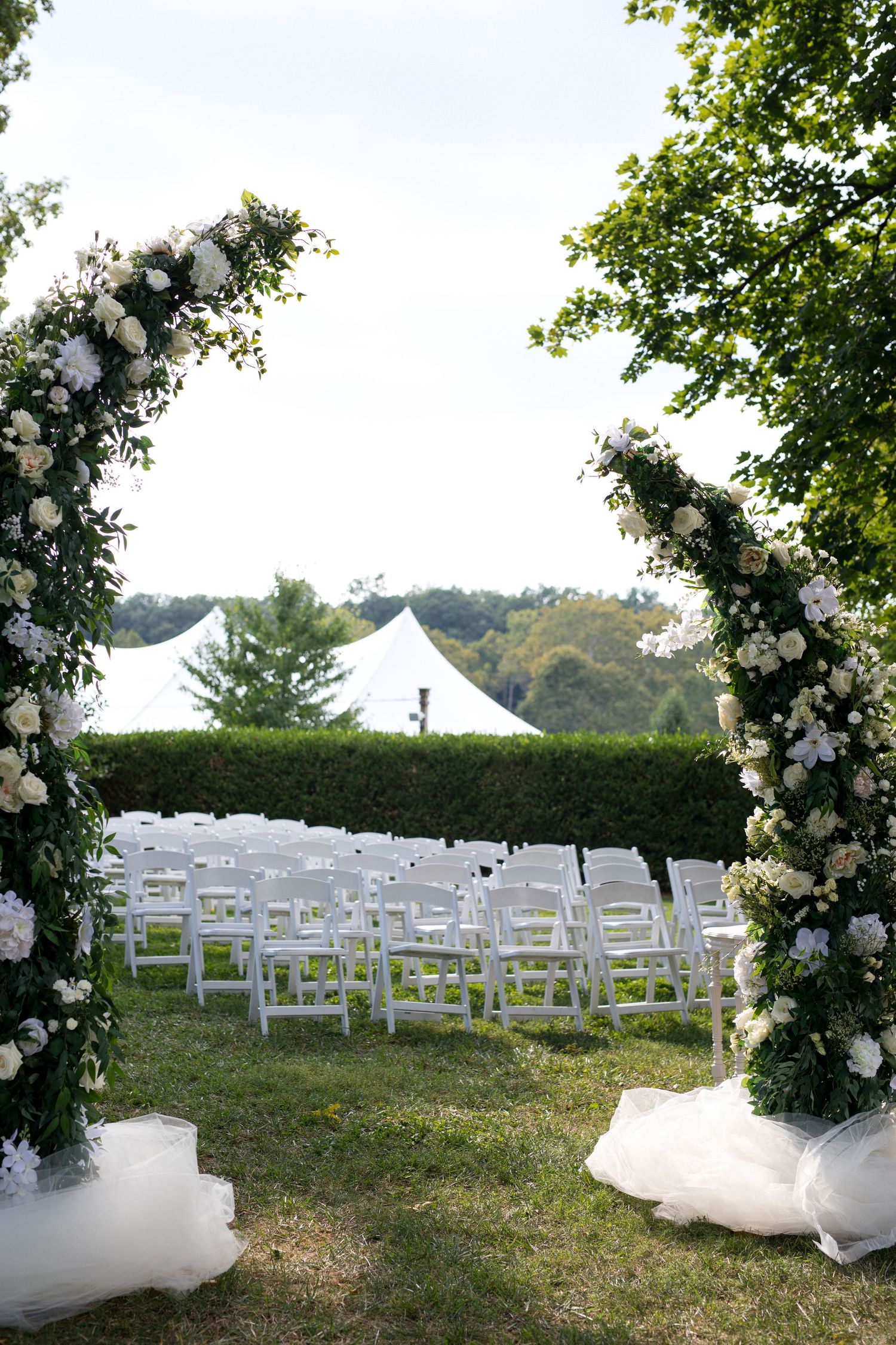 Elegant outdoor wedding ceremony setup with white chairs and floral arch decorations in a garden setting.