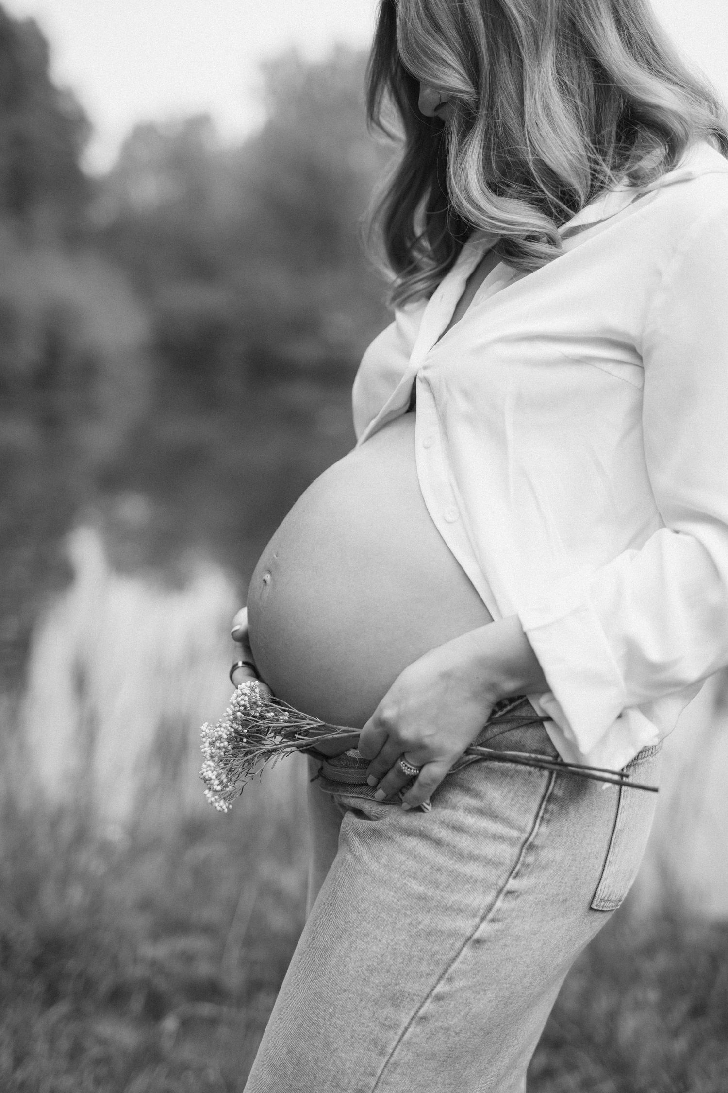 Black and white photo of pregnant woman outdoors holding wildflowers, showing baby bump in white shirt and jeans.