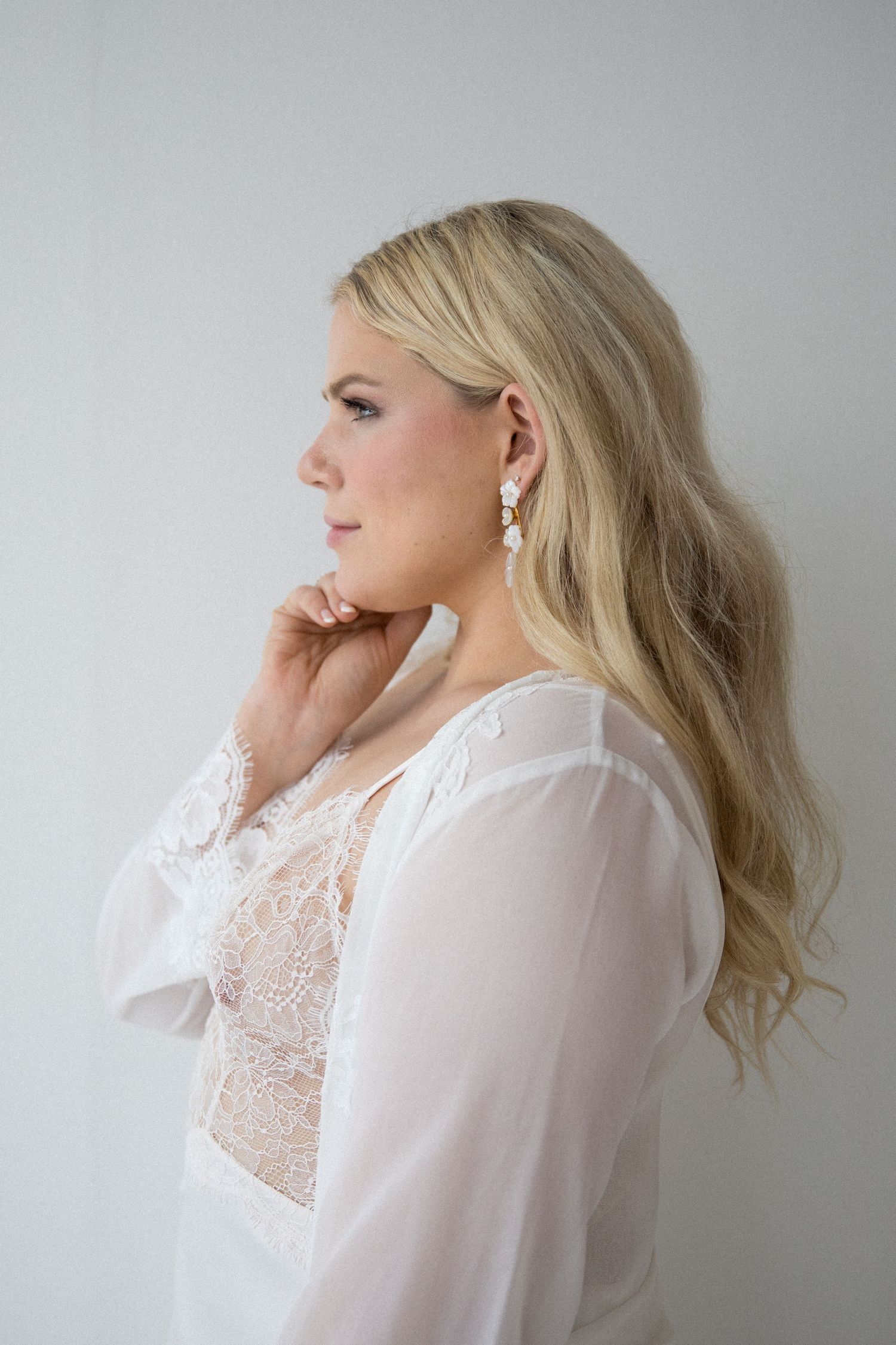 Blonde woman in white lace bridal robe with elegant drop earrings, posing in soft profile view against light background.