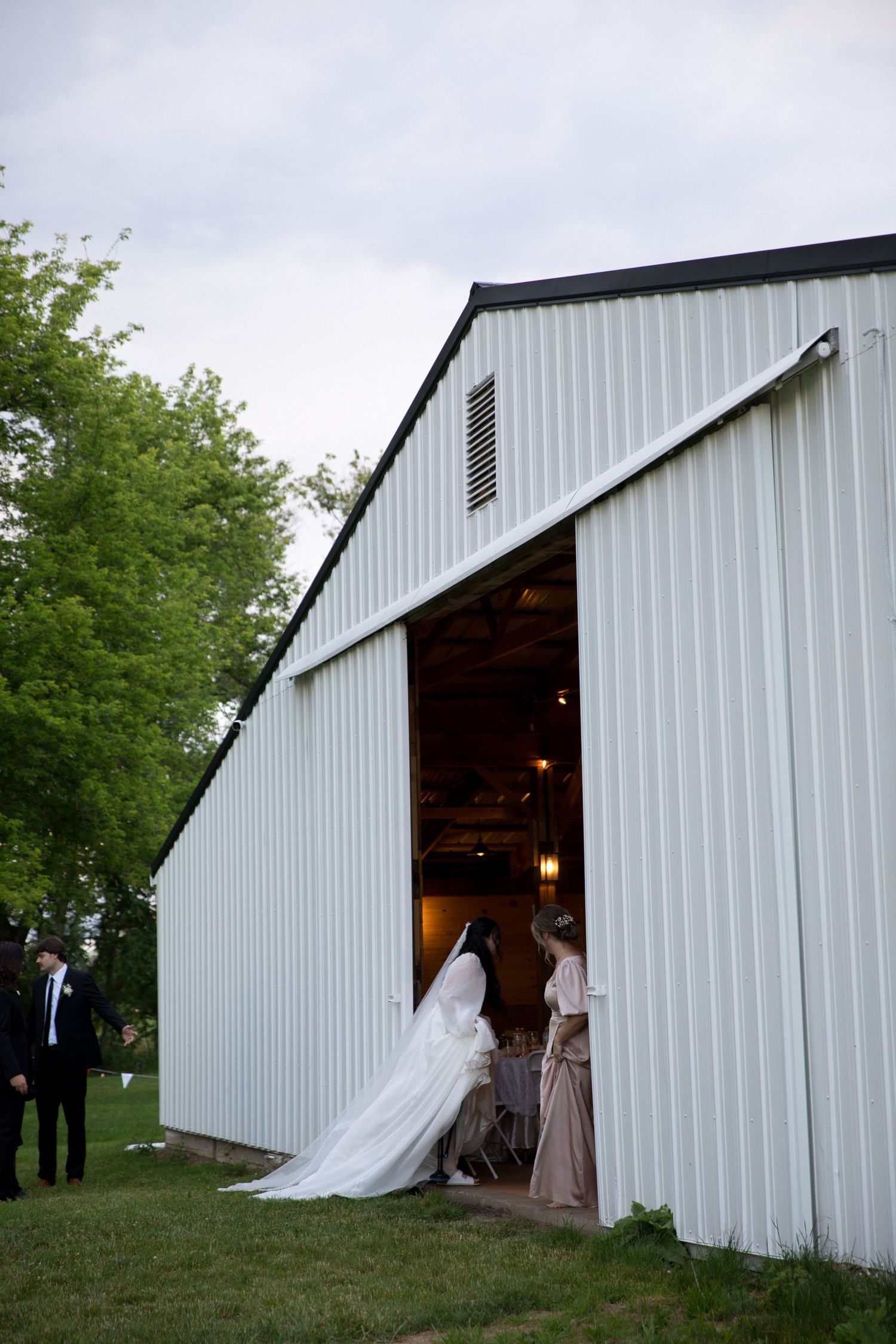 Bride in white gown entering a white barn venue with bridesmaids at a rustic outdoor wedding reception at dusk.