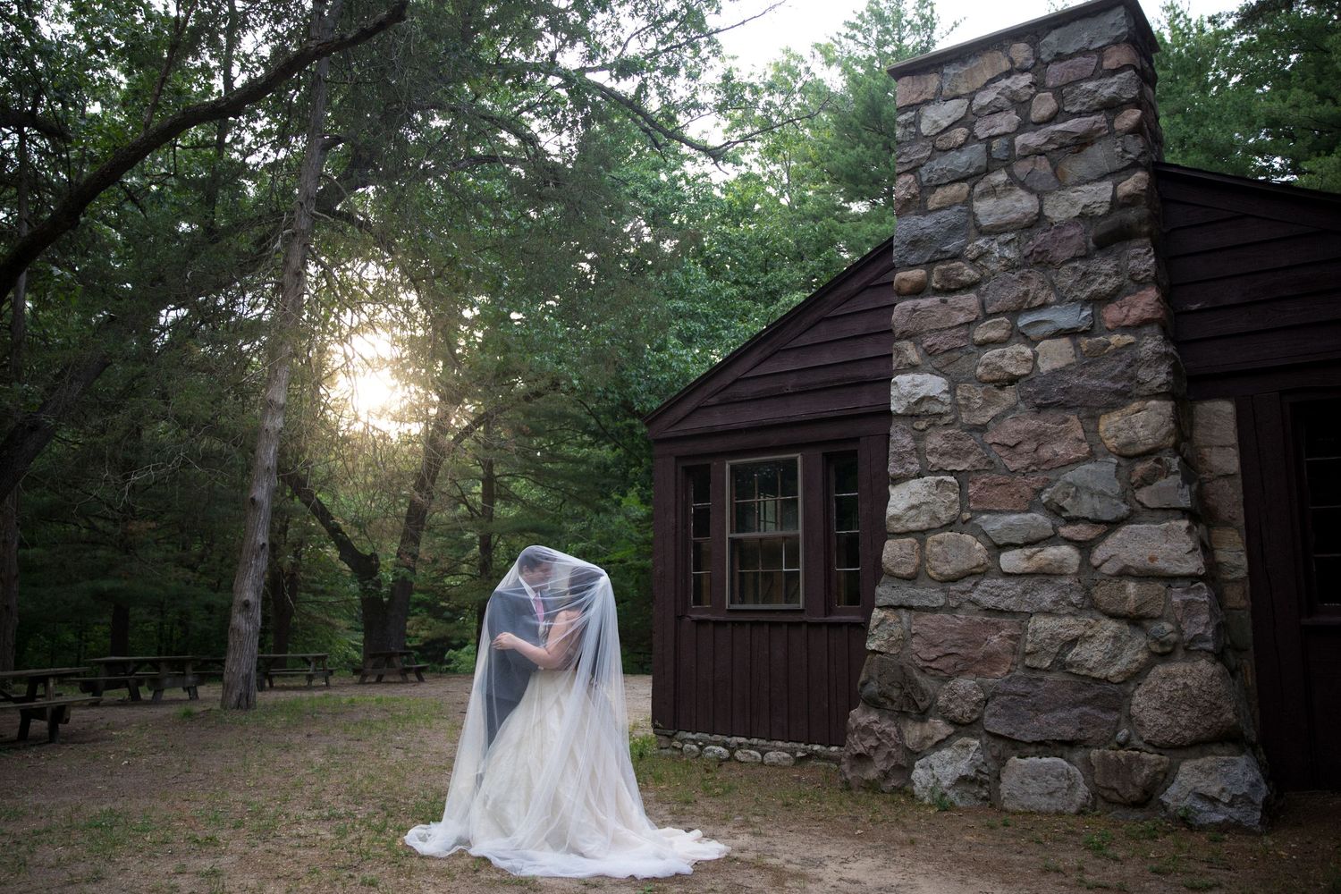 Bride in white wedding gown and veil stands before rustic stone cabin surrounded by trees at sunset.
