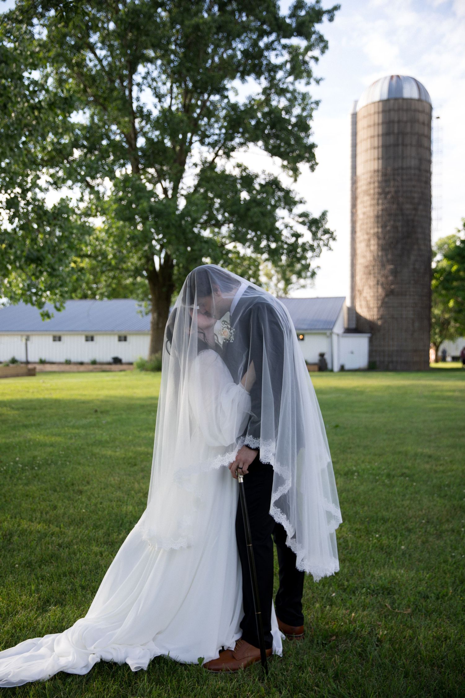 Bride and groom sharing a kiss under a flowing veil on a green lawn with a rustic barn and silo backdrop.
