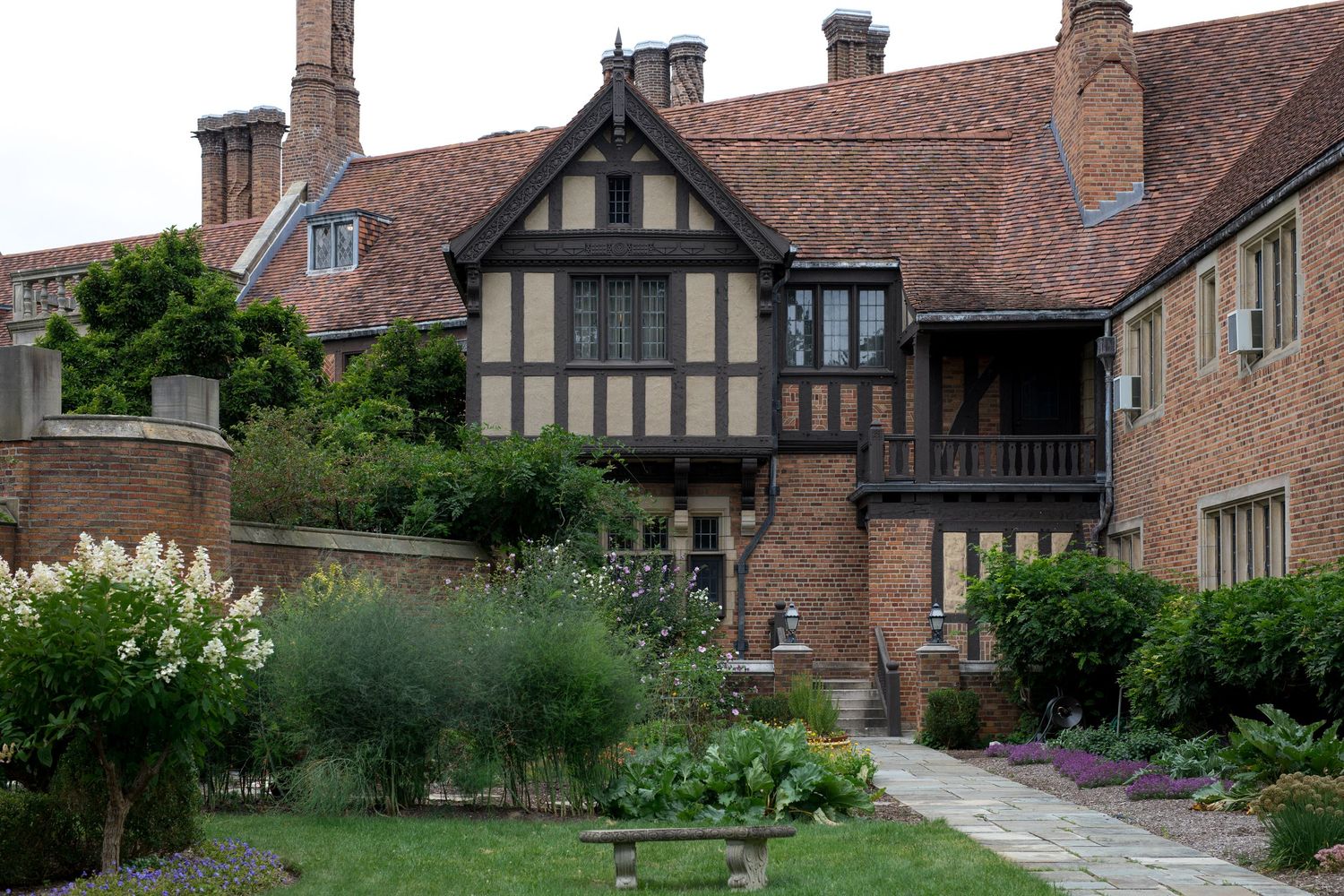 Tudor-style brick manor house with timber framing, balcony, and lush cottage garden with flowering plants and manicured lawn.