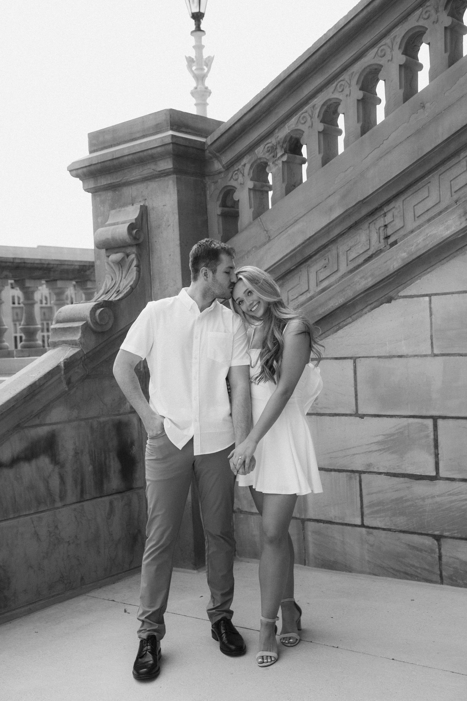 Black and white photo of couple holding hands and kissing on stone building terrace.
