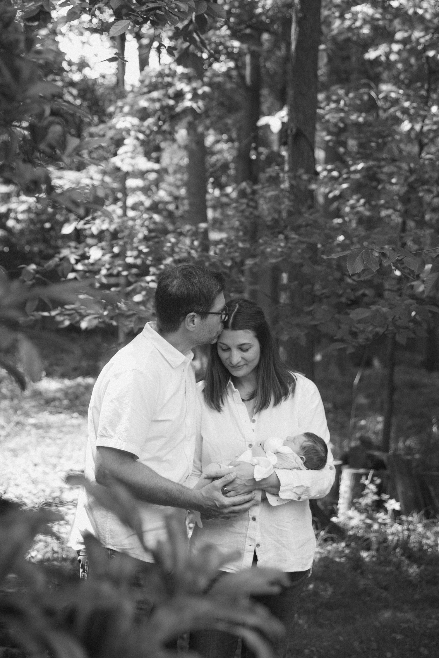 Black and white photo of parents lovingly gazing at their newborn baby outdoors in a lush green forest setting.