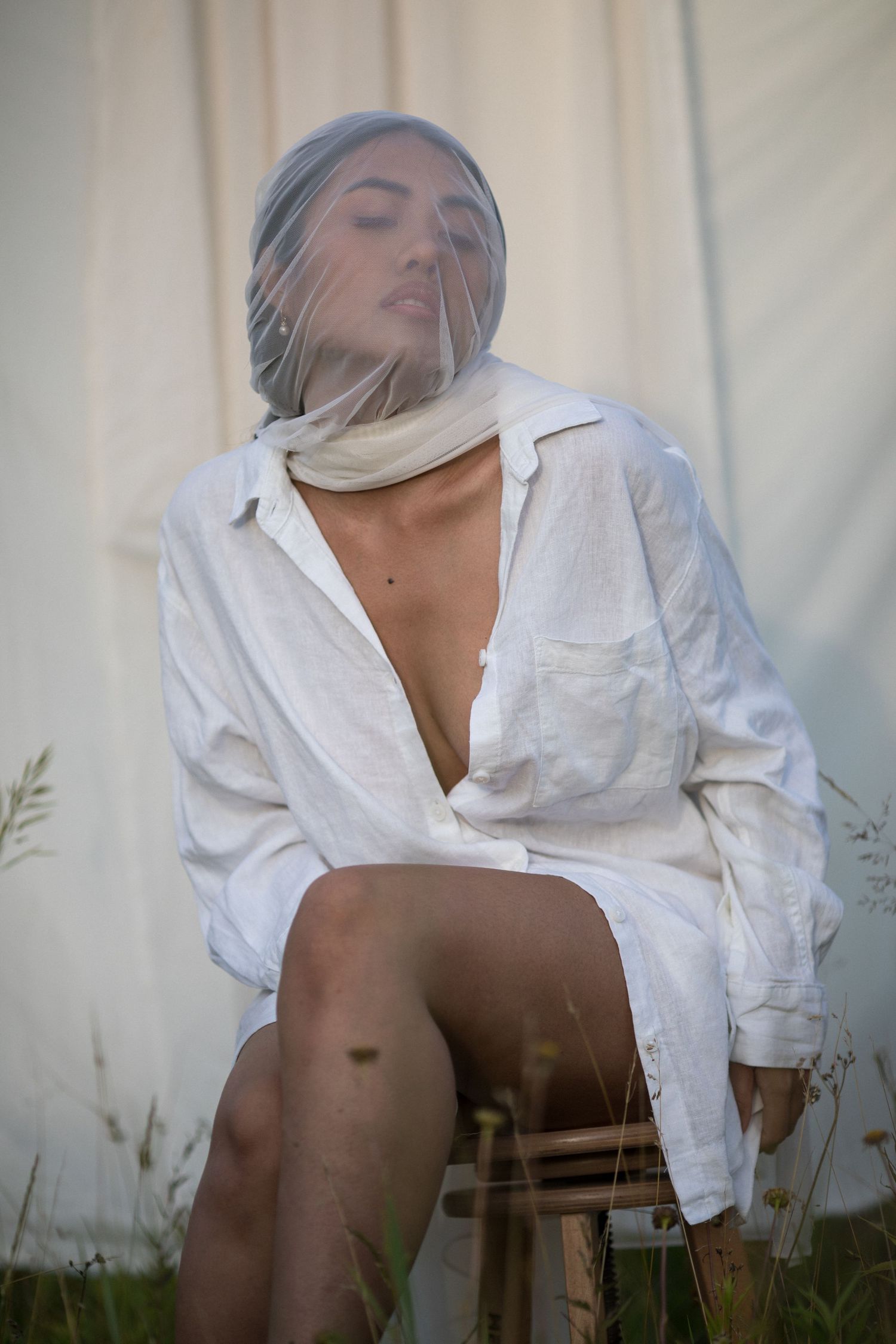 A person in a white shirt sits on a wooden chair against a light curtain backdrop.