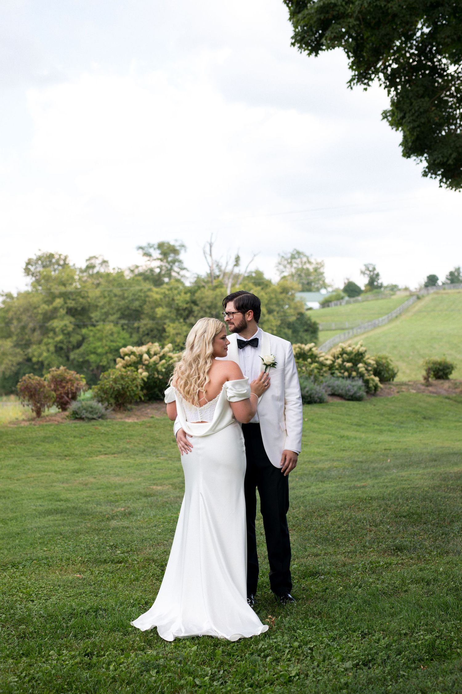 A romantic couple in wedding attire embrace while walking on a grassy hillside during a sunny outdoor celebration.