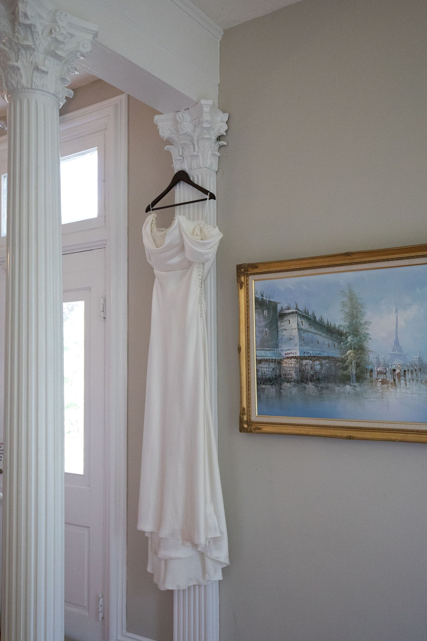 White wedding dress hanging on a hanger against a wall with decorative columns and framed artwork.