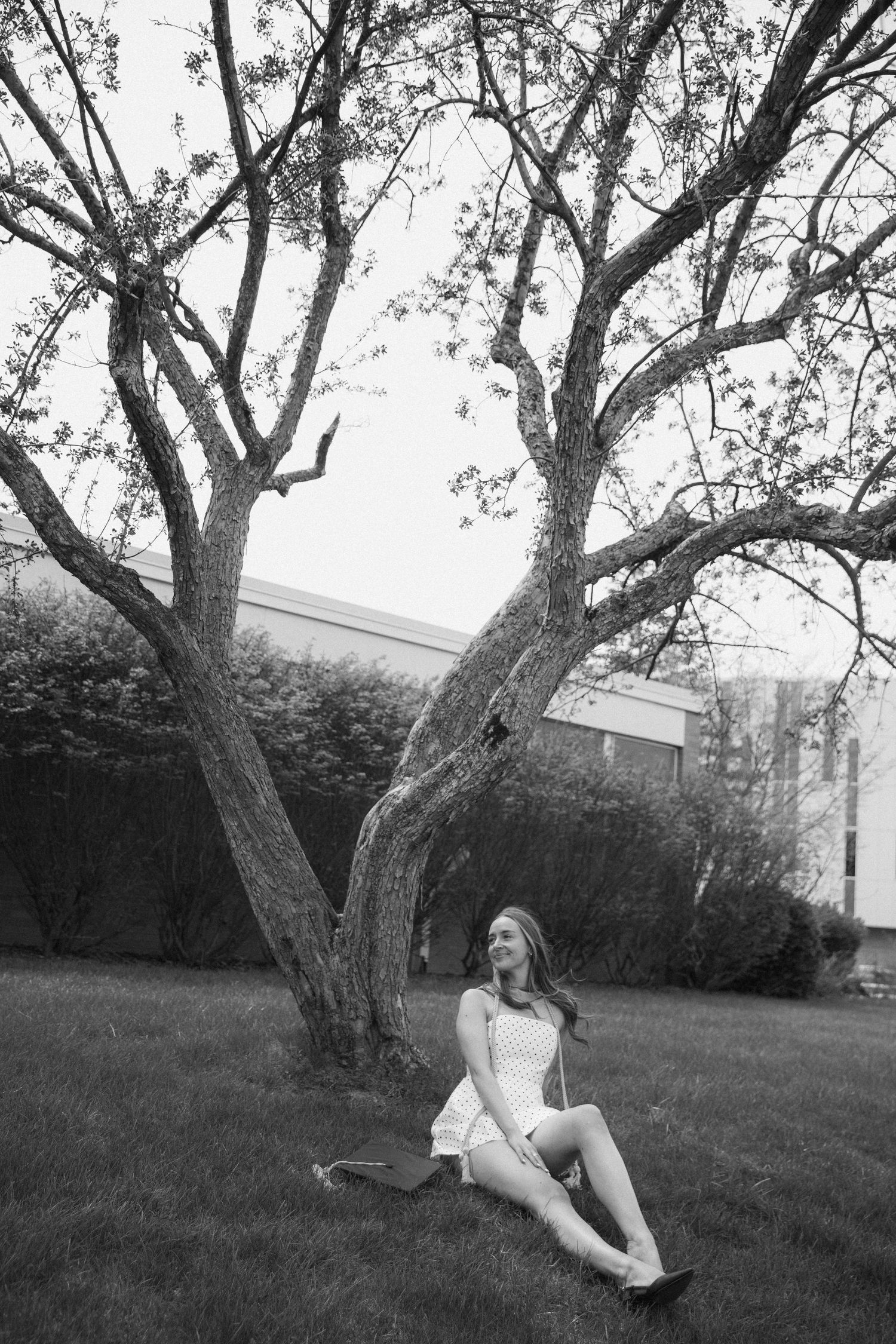Black and white photo of a young woman in a white dress sitting under a large tree on a green lawn.
