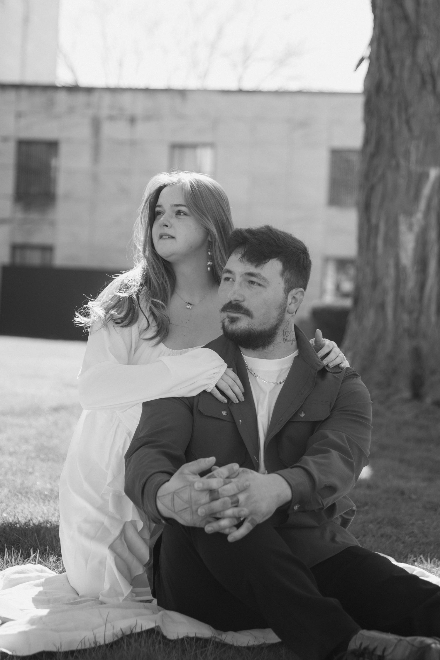 Black and white photo of a couple sitting outdoors, woman in white dress embracing man seated on grass near a tree.