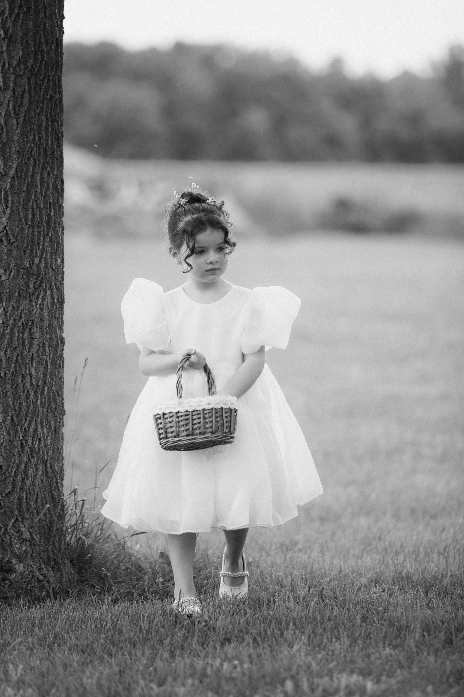 Black and white photo of a young flower girl in a white dress holding a wicker basket near a tree.