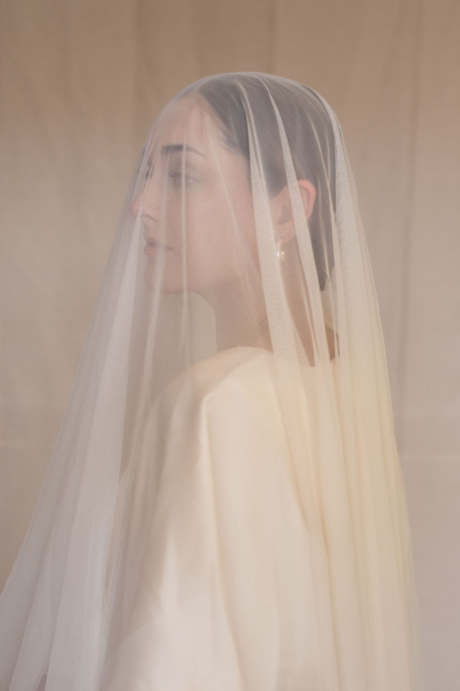 A bride wearing an ethereal sheer veil poses in soft lighting against a neutral background, creating a dreamy bridal portrait.