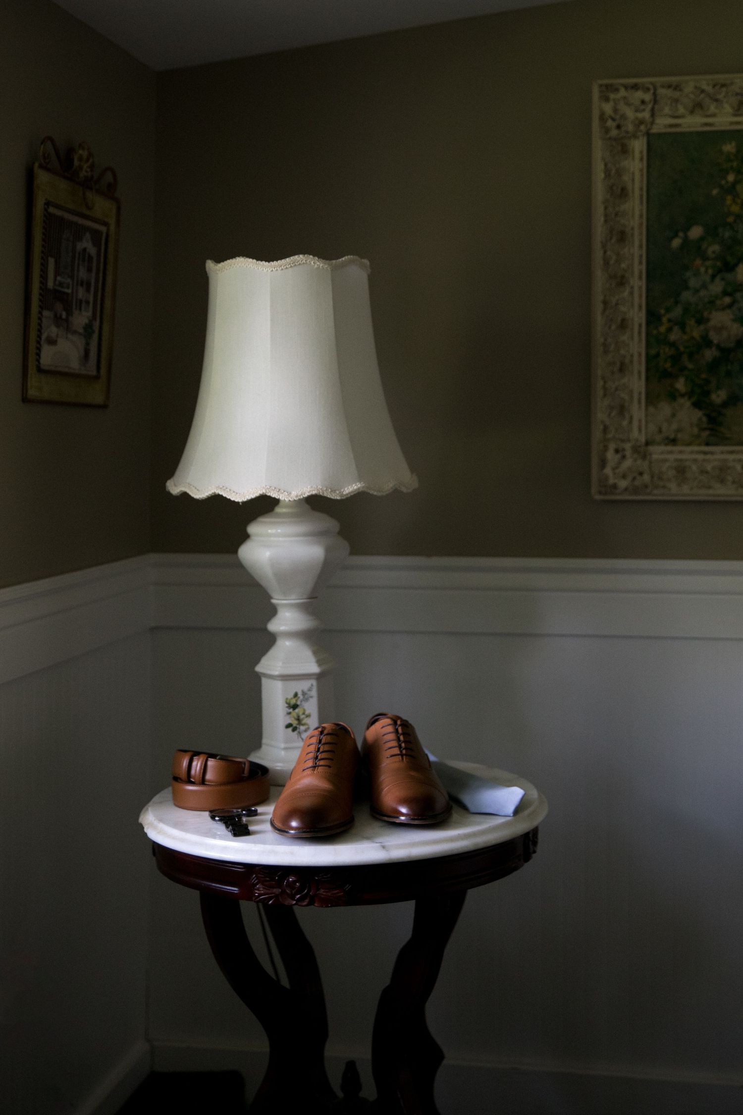 A vintage white table lamp sits atop a dark wooden table with leather football equipment in a dimly lit room.