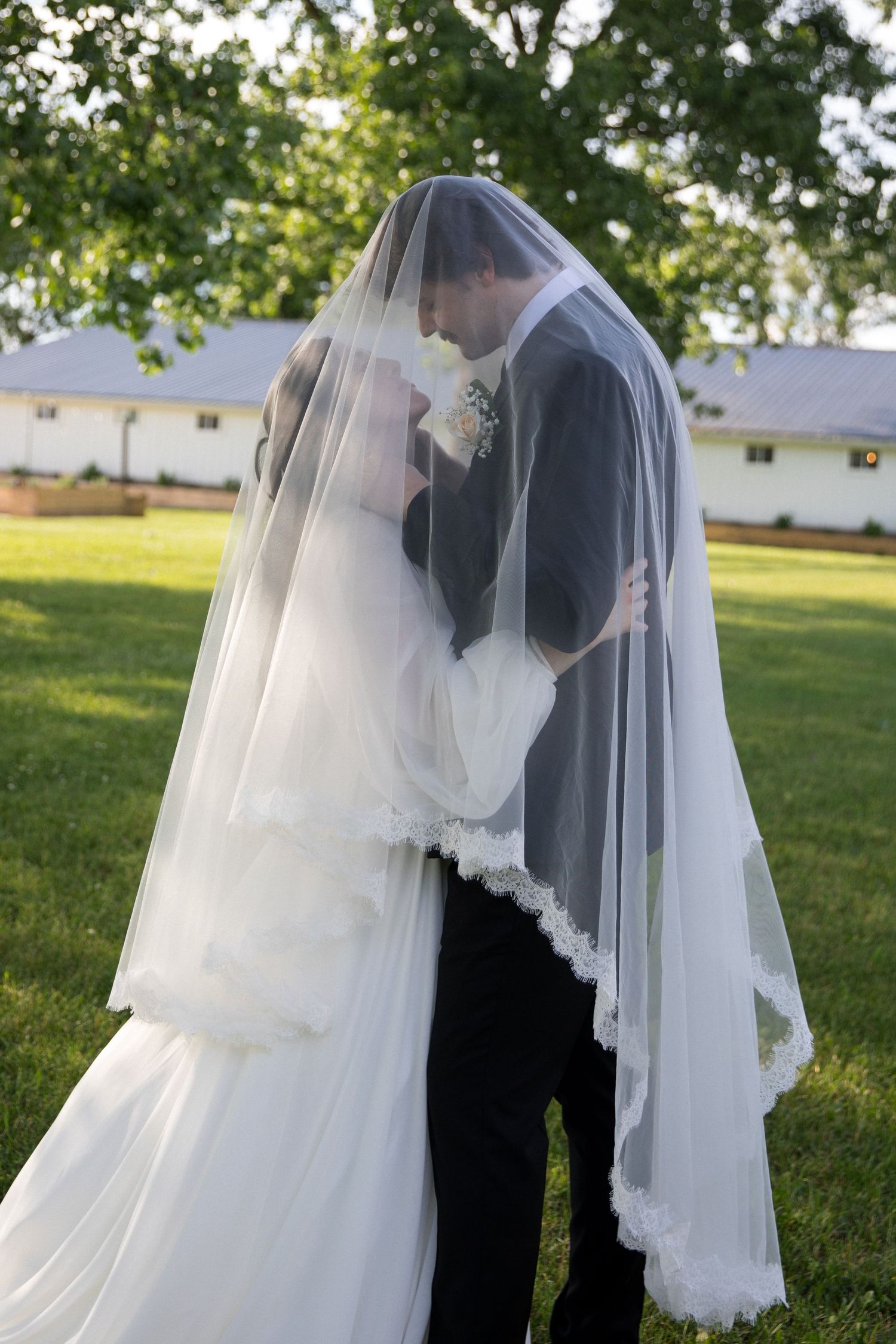 Bride and groom sharing a romantic kiss under a flowing white wedding veil outdoors on green grass.