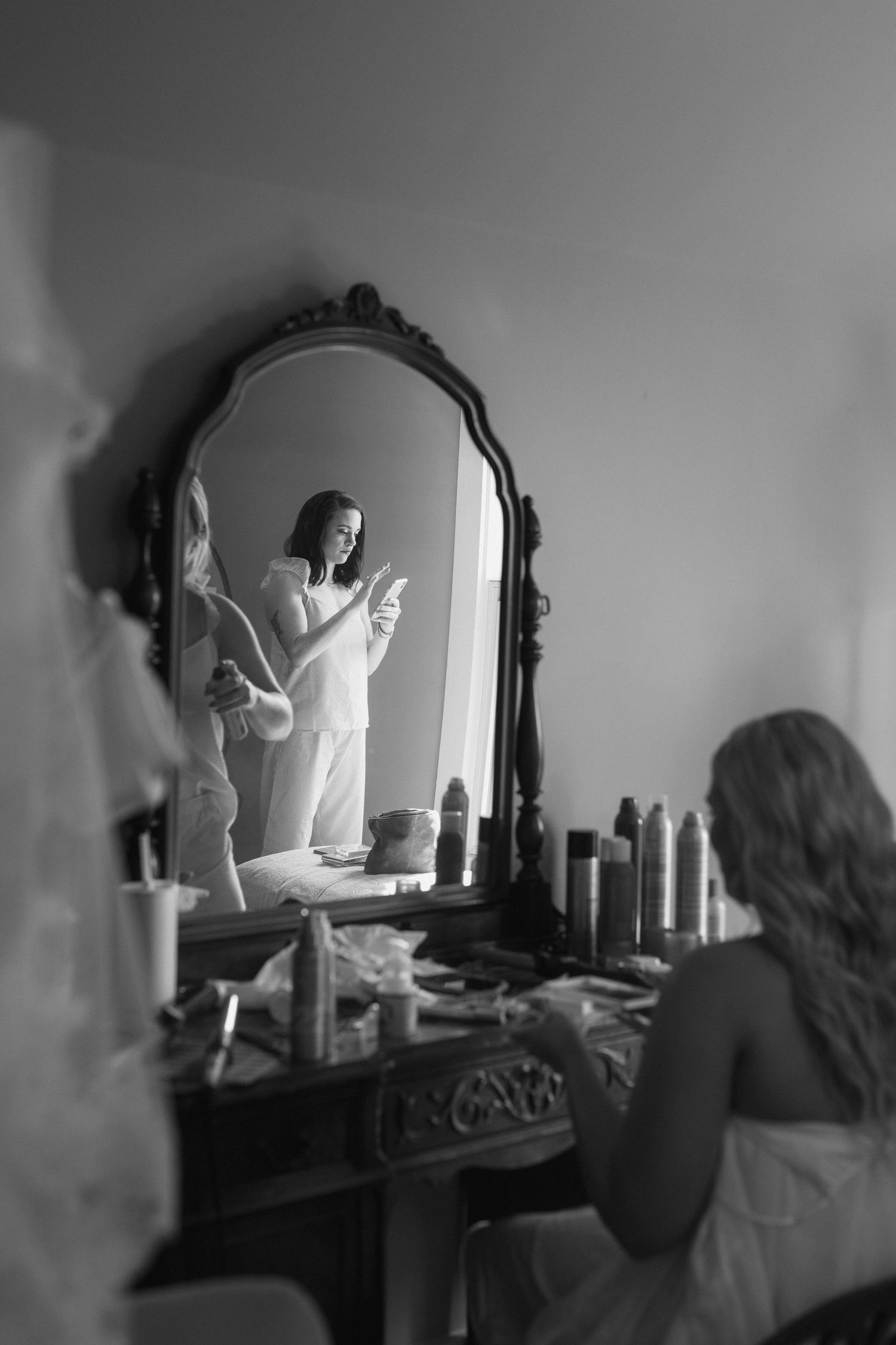 Black and white photo showing a person getting ready at a vintage vanity mirror surrounded by beauty products.