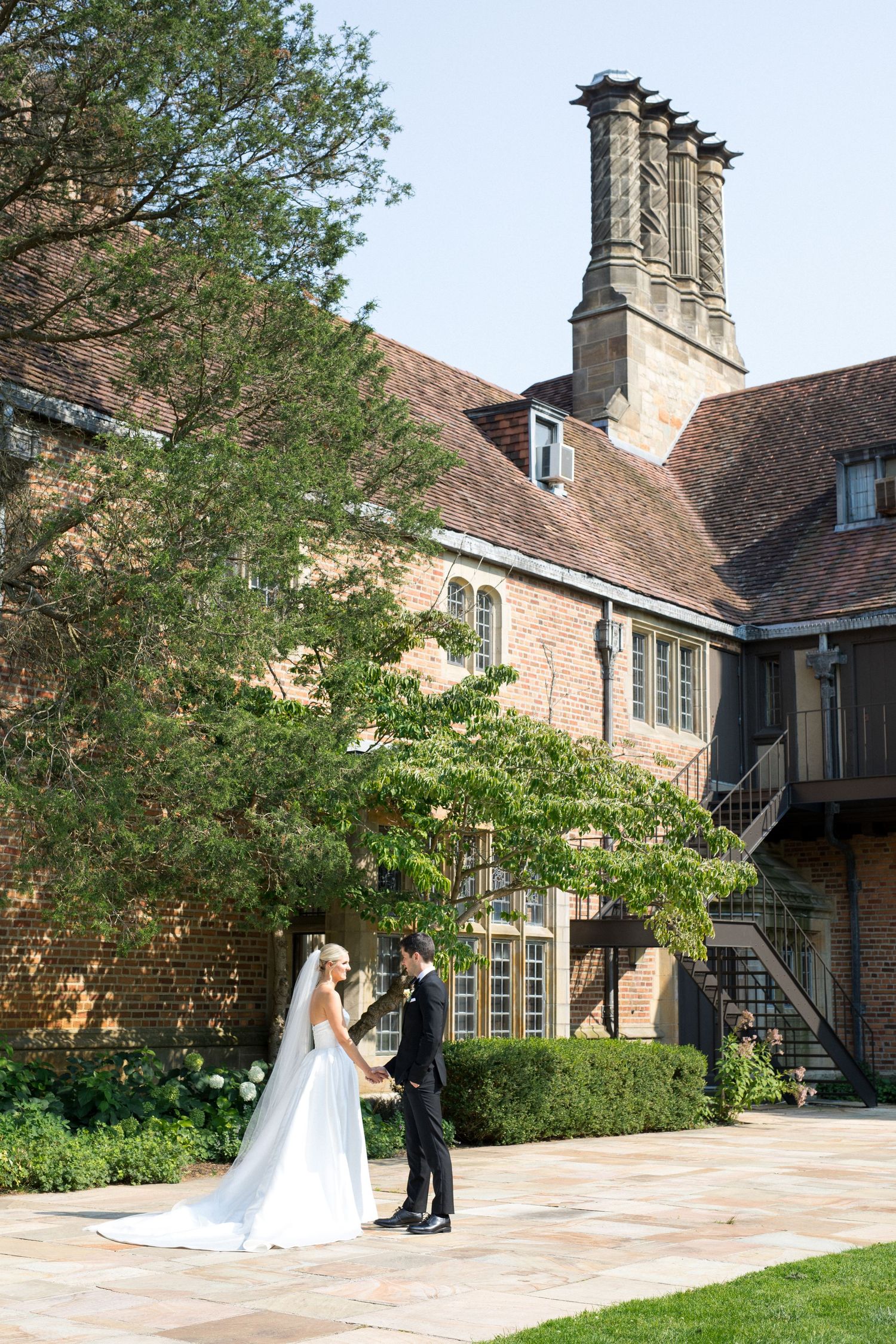 A wedding couple poses for photos on a stone patio outside a traditional brick manor house with lush gardens.