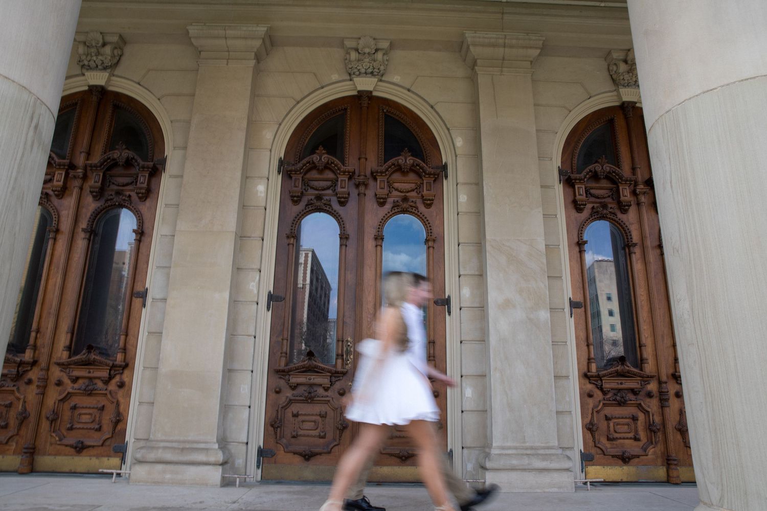 A woman in a white dress walking past ornate wooden arched doors of a historic building.