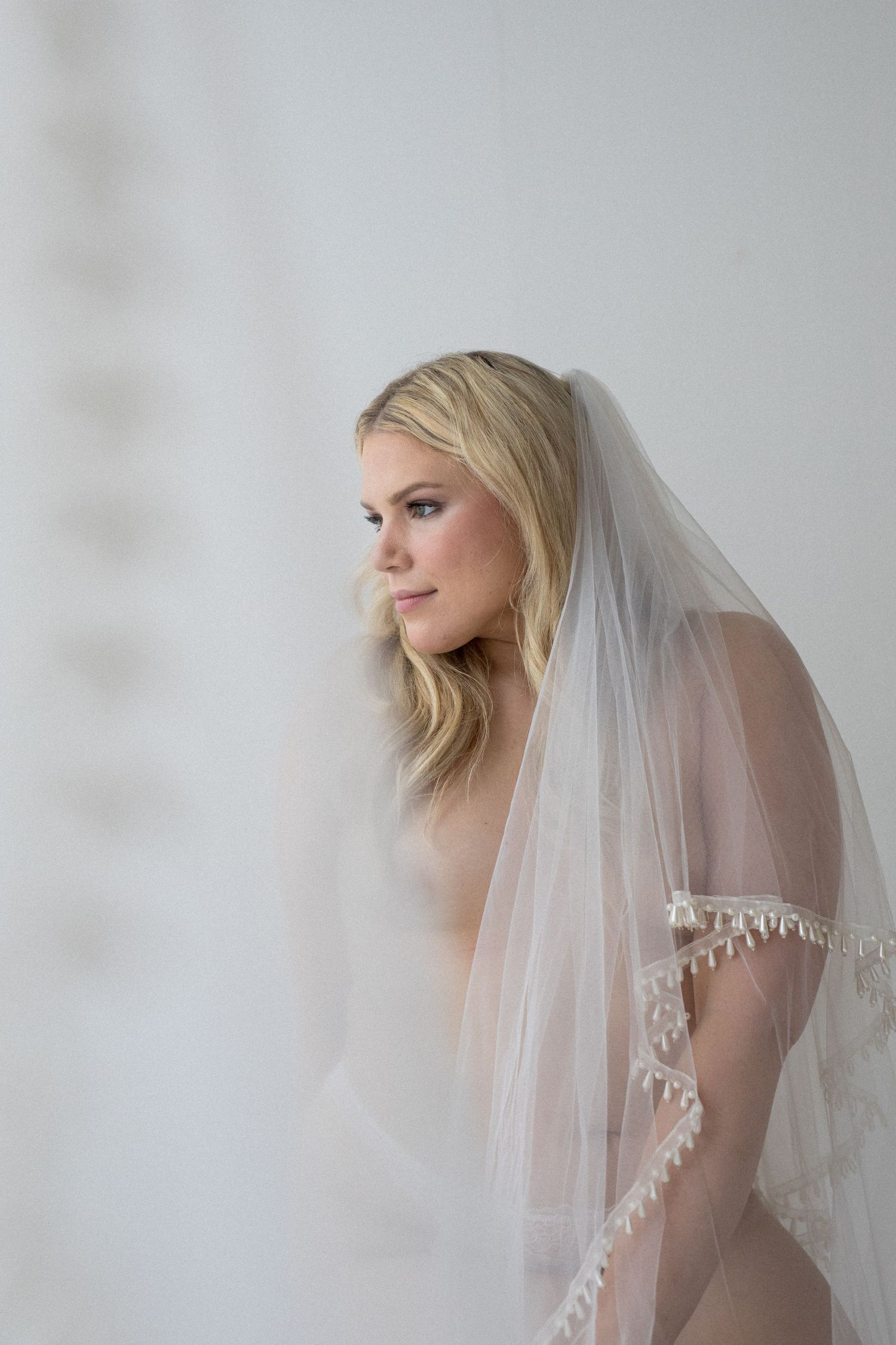 Bride wearing a delicate lace-trimmed wedding veil, posing elegantly against a soft white background.
