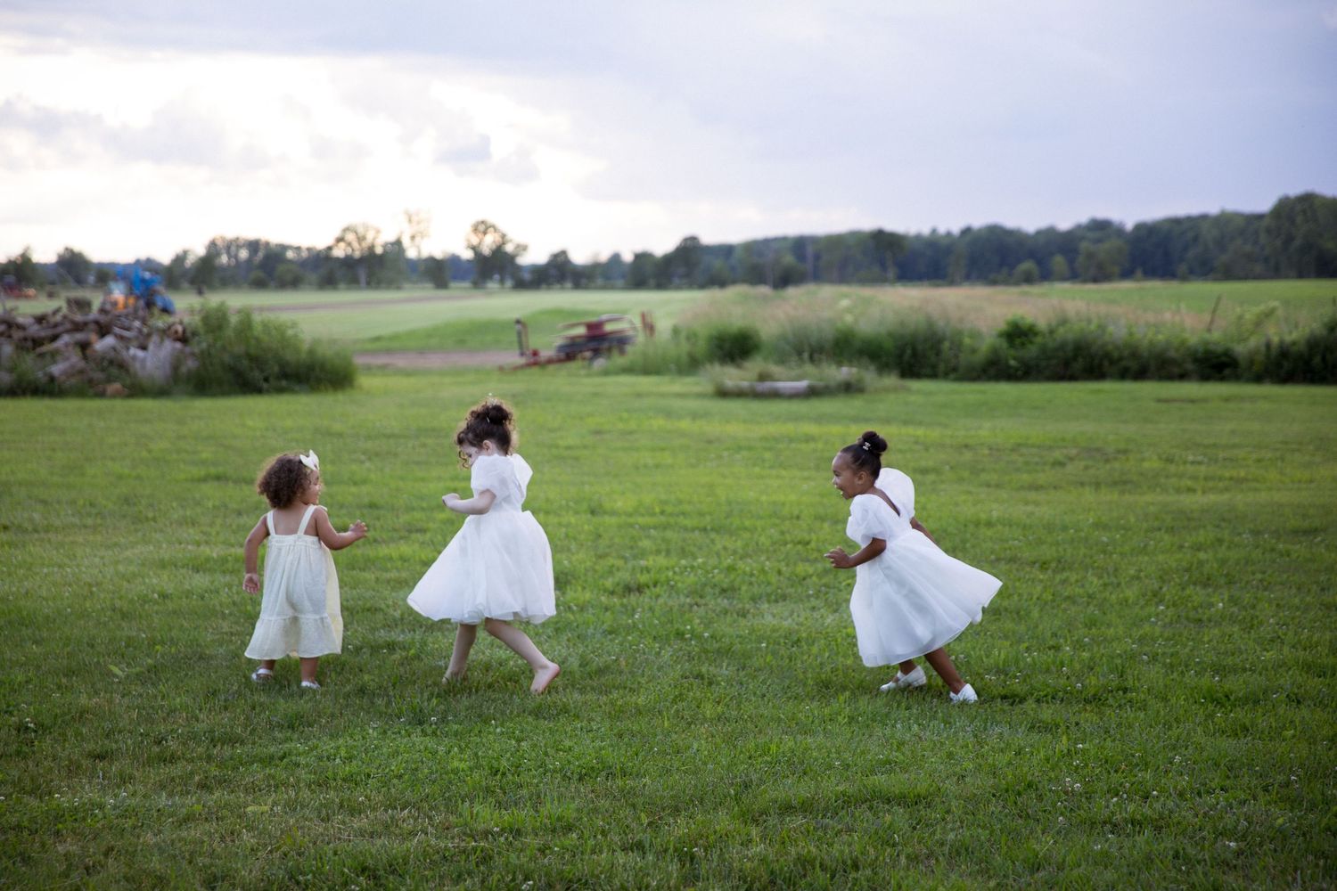 Three young girls in white dresses running and playing on a lush green field at dusk.