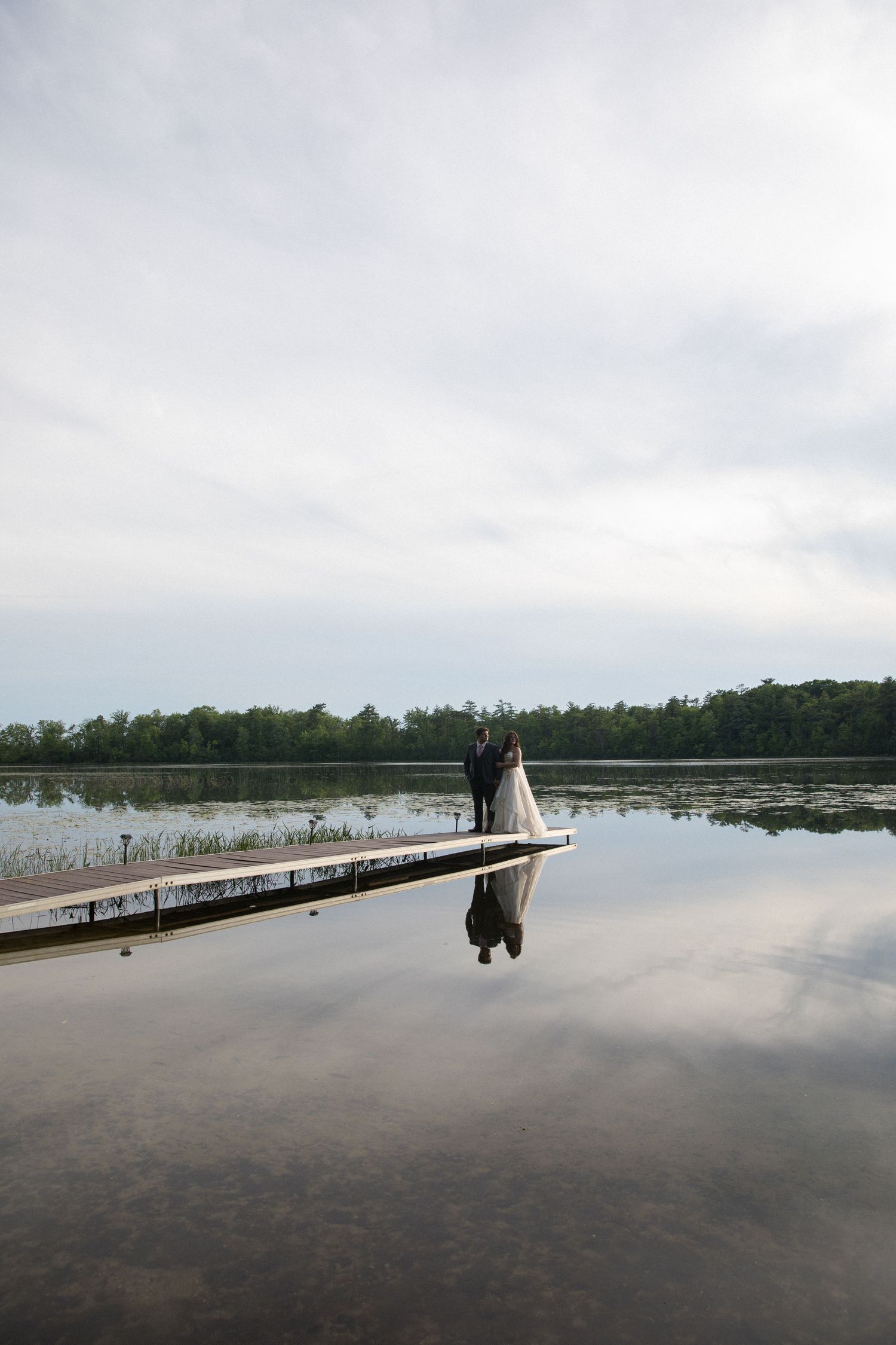 Bride and groom stand on a dock reflecting on a calm lake, surrounded by lush trees under a cloudy sky.