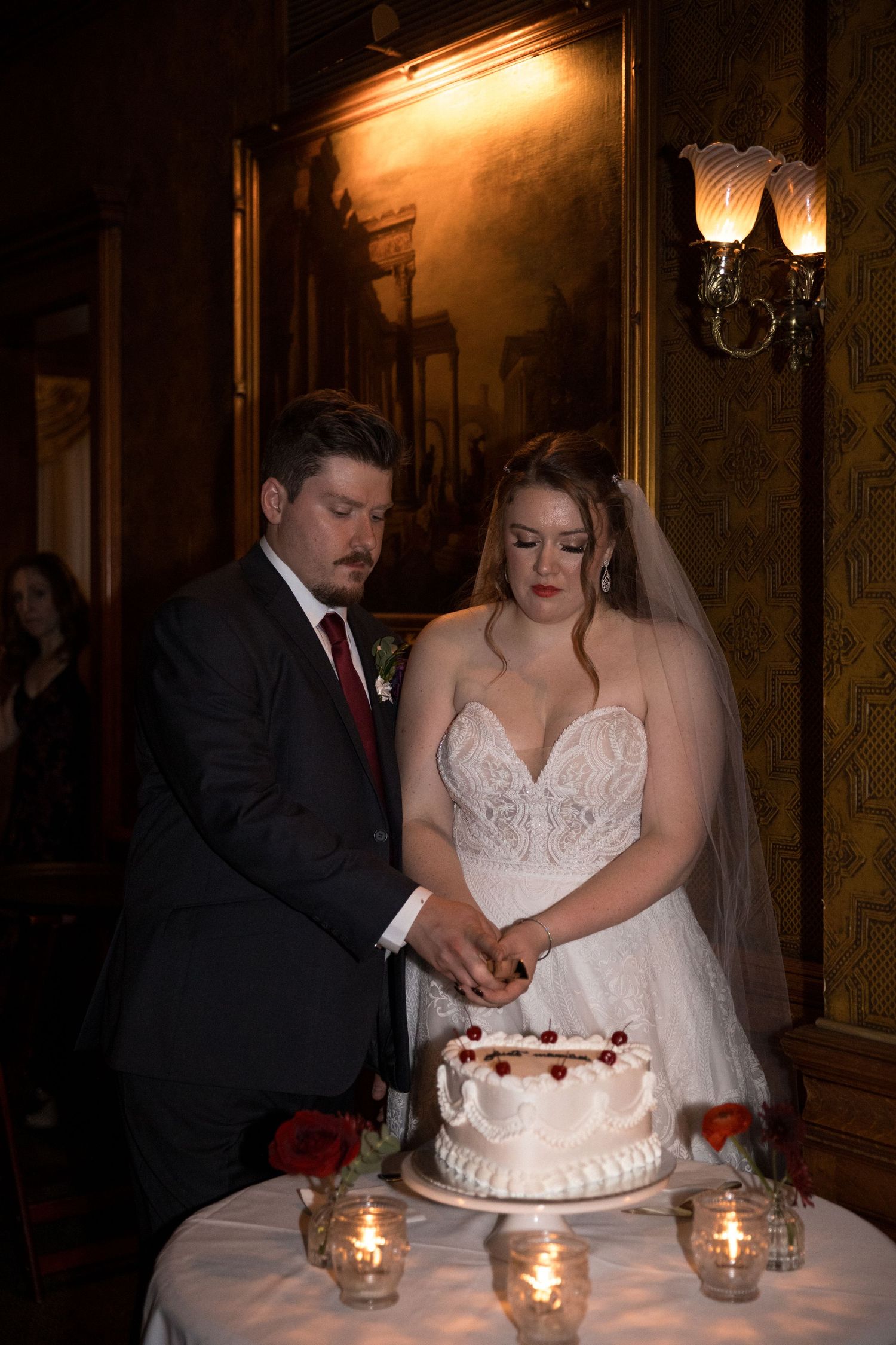 A couple in formal wedding attire cuts their white wedding cake together at an elegant candlelit reception table.