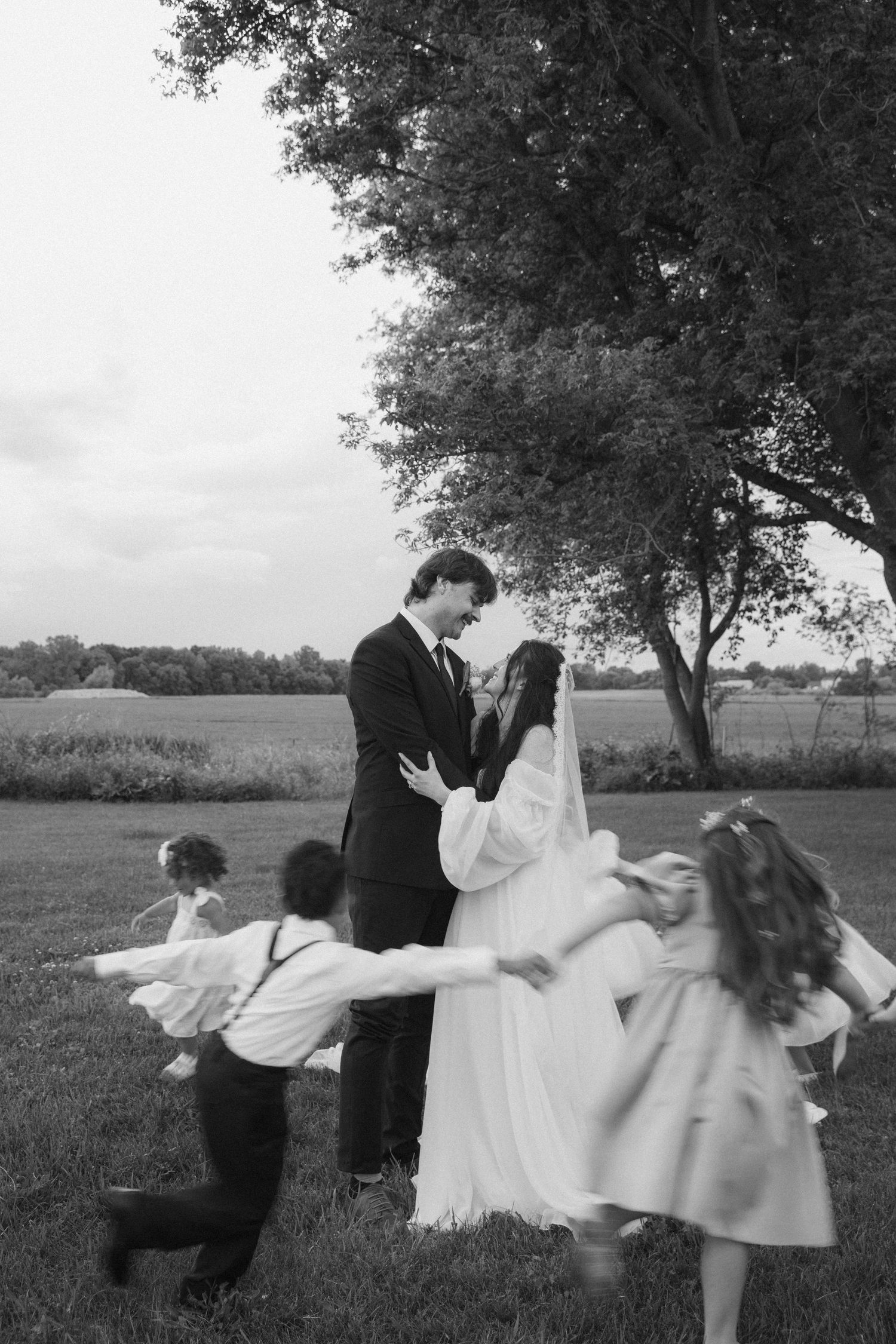 Black and white photo of a bride and groom surrounded by children playing outdoors near a large tree.