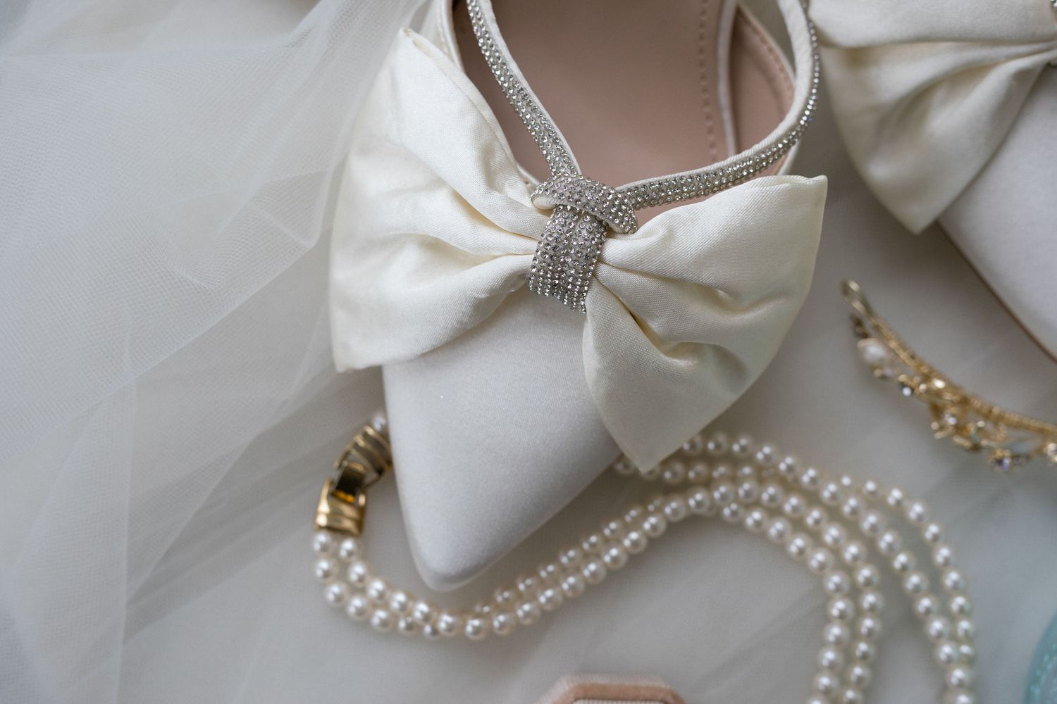 Elegant white bridal shoe with a large bow accent displayed alongside delicate pearl necklaces and tulle fabric.