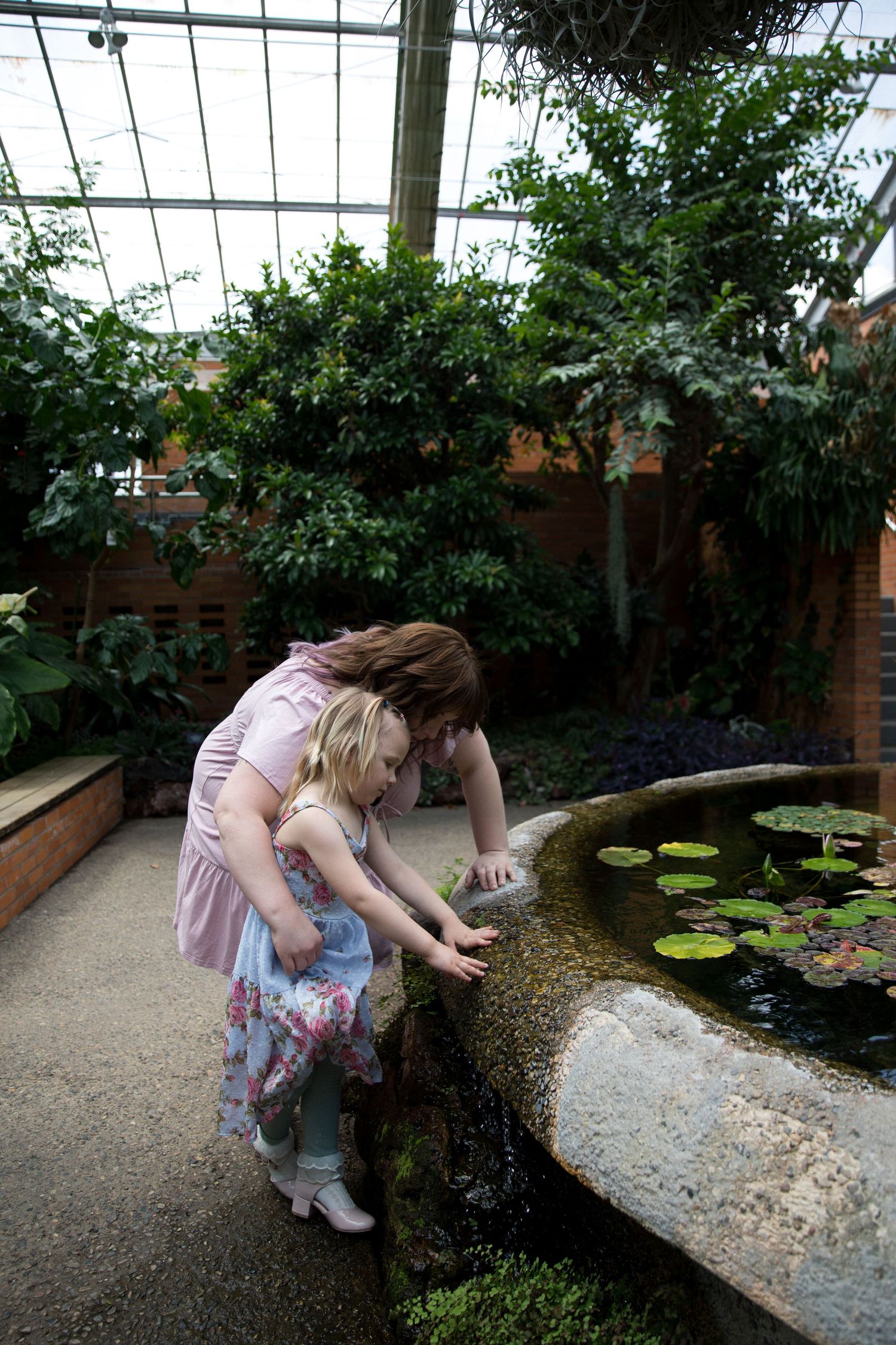 Woman and young girl leaning over a lily pond in a lush greenhouse conservatory.