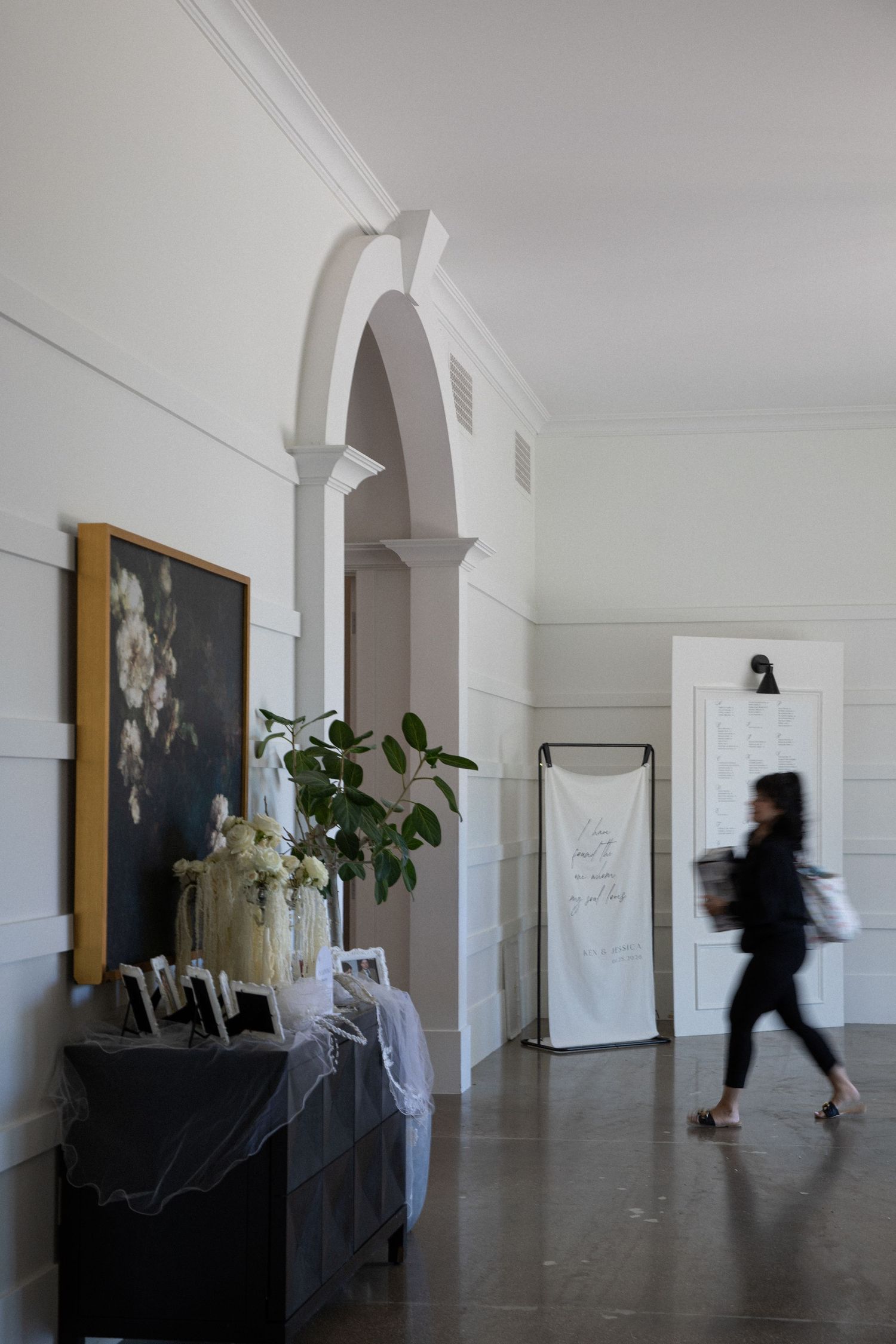 Elegant wedding venue hallway with arched doorway, floral arrangements, and a blurred guest walking past decorative signage.