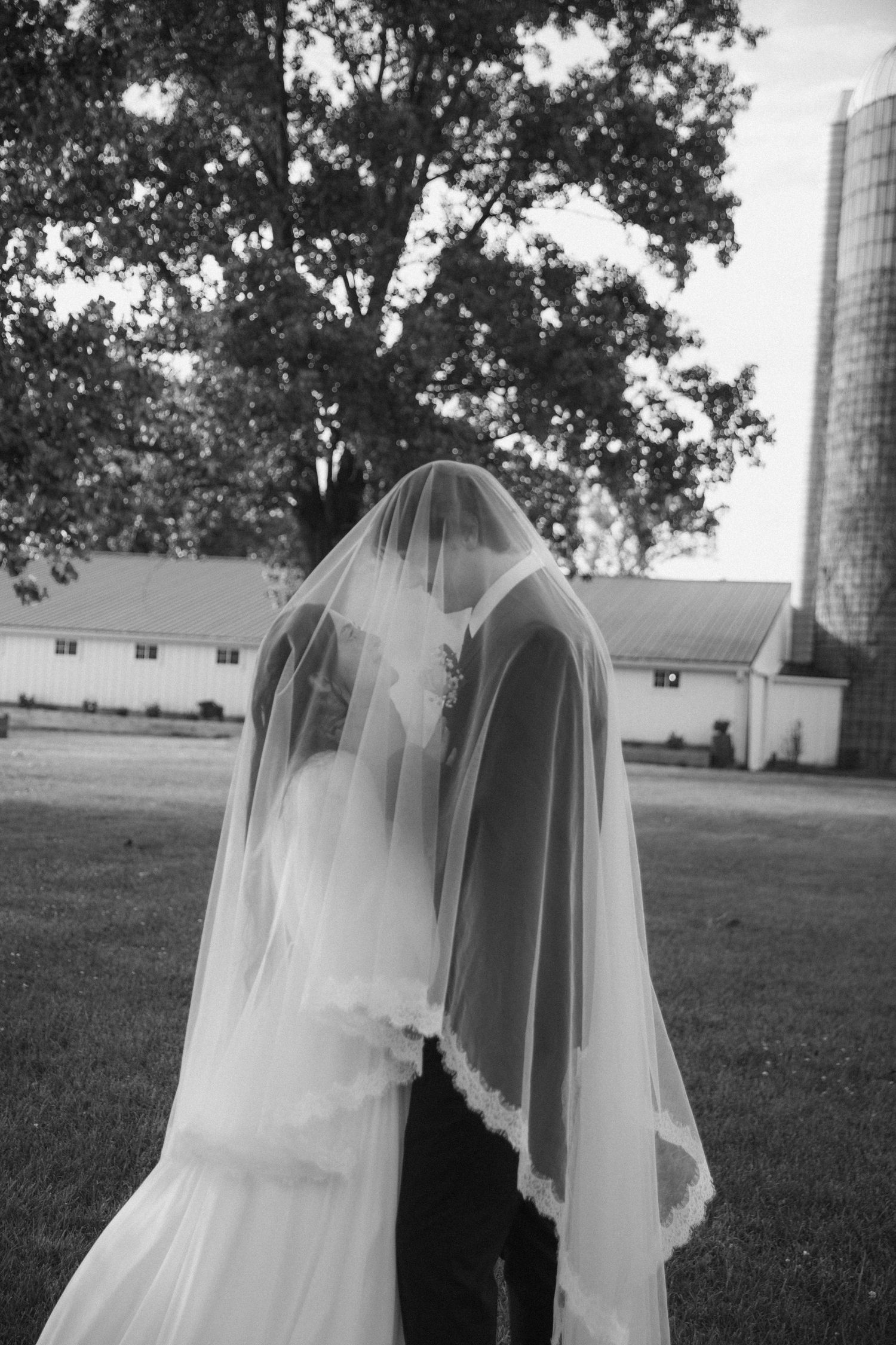 Black and white photo of a bride and groom sharing a kiss hidden beneath a flowing wedding veil outdoors.