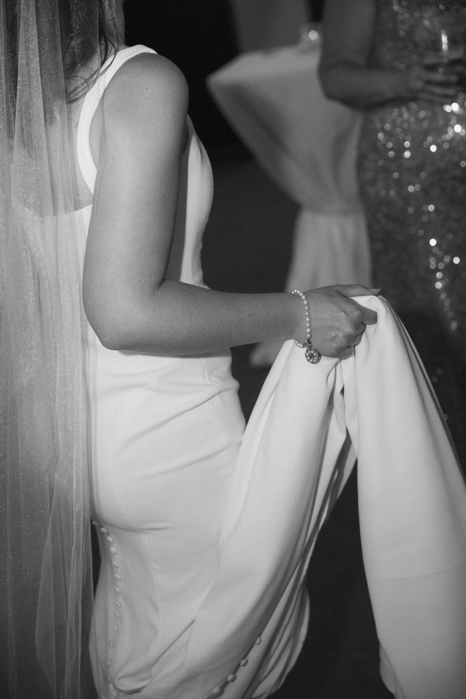 Black and white photo of a bride in a sleek white gown holding her dress train, wearing a pearl bracelet and veil.