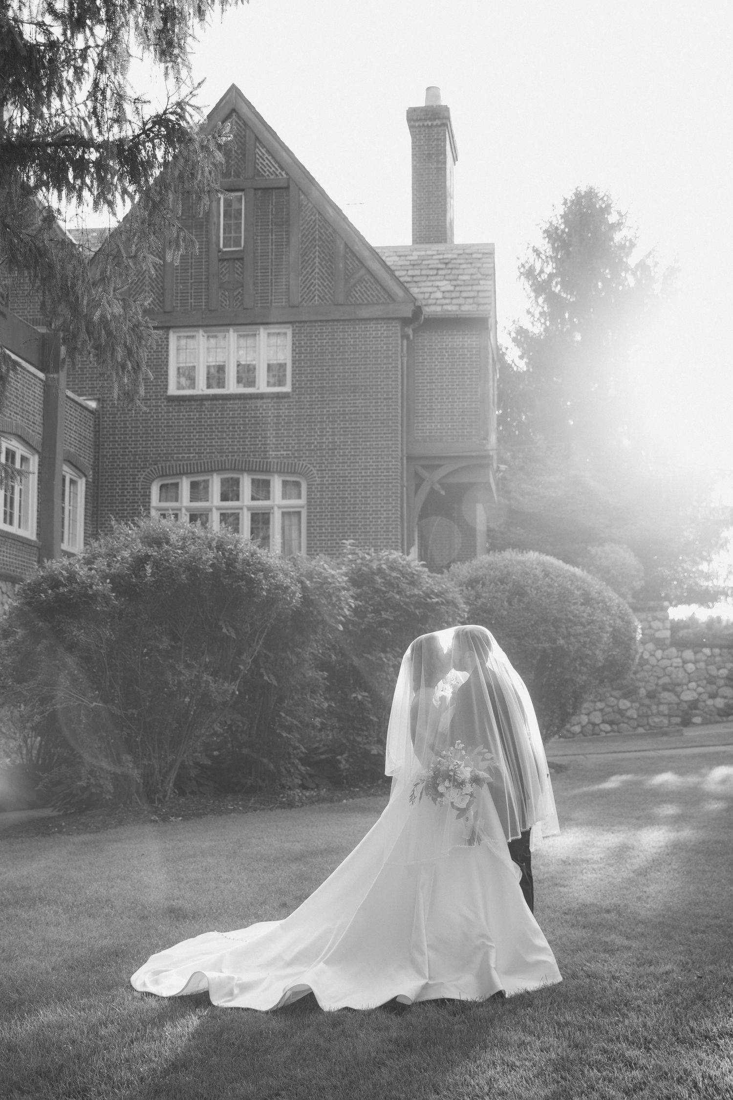 A bride in a flowing white wedding gown and veil stands on a lawn in front of a Victorian-style house in black and white.