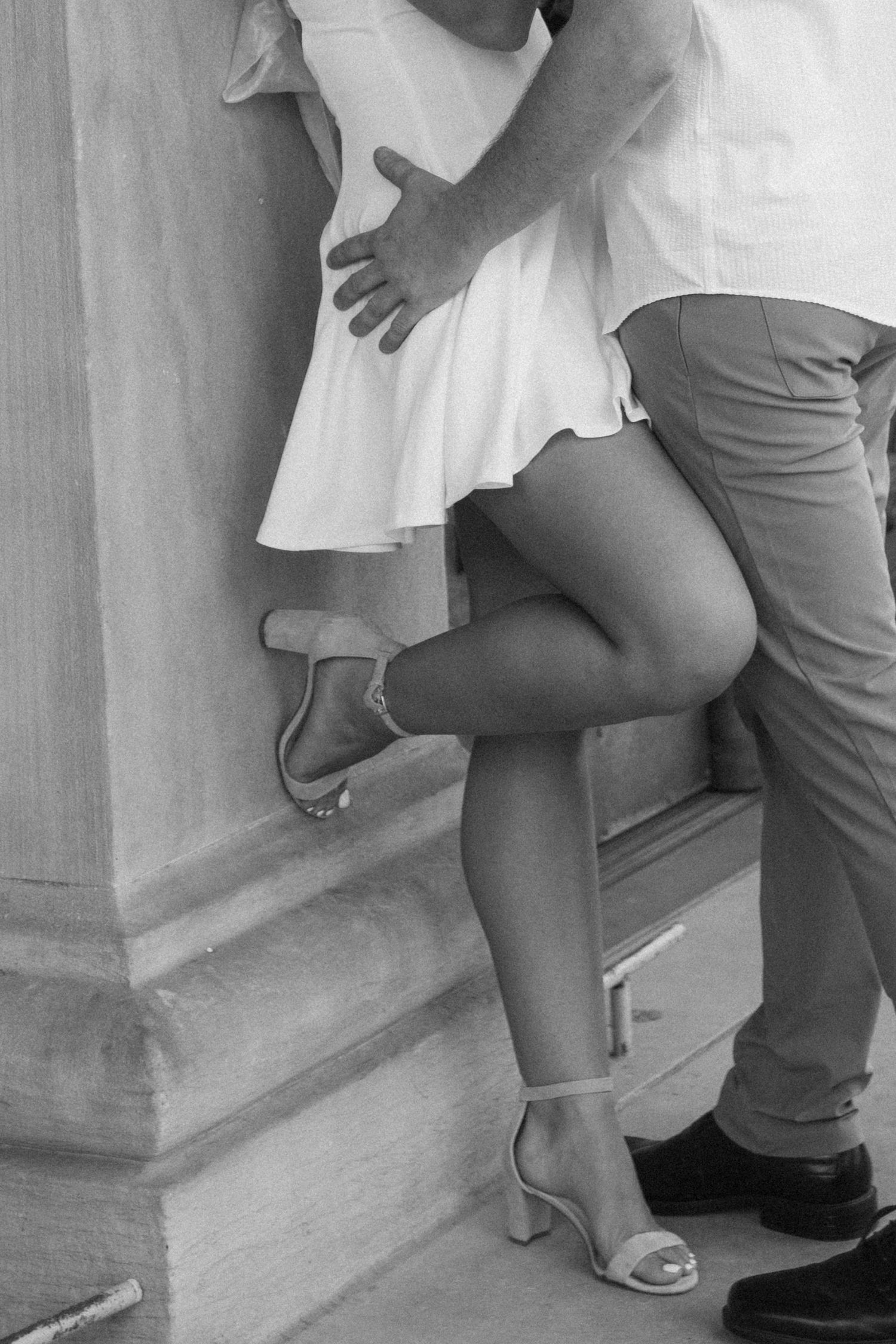 Black and white close-up of couple embracing by stone column, woman in white dress and heeled sandals.