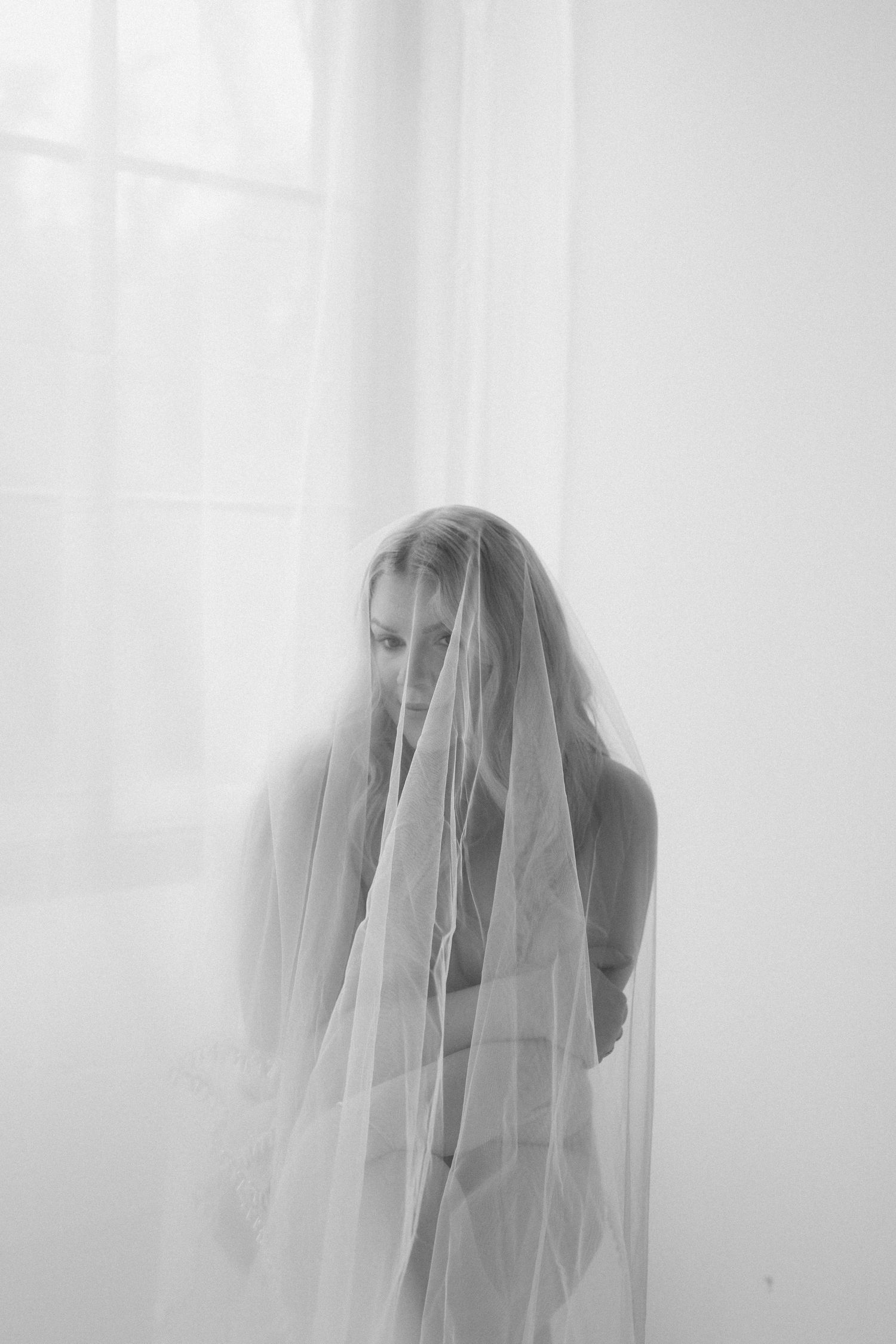 Black and white photo of a bride draped in a sheer wedding veil, standing in soft natural light.