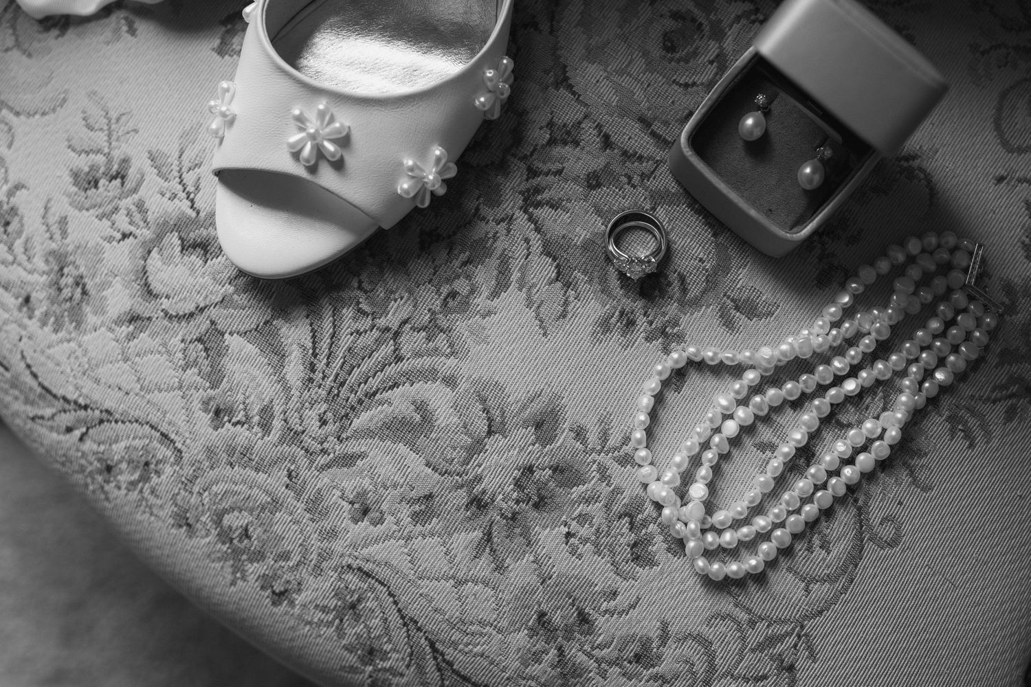 Wedding accessories including pearls, rings, and bridal shoes arranged on lace fabric in black and white.