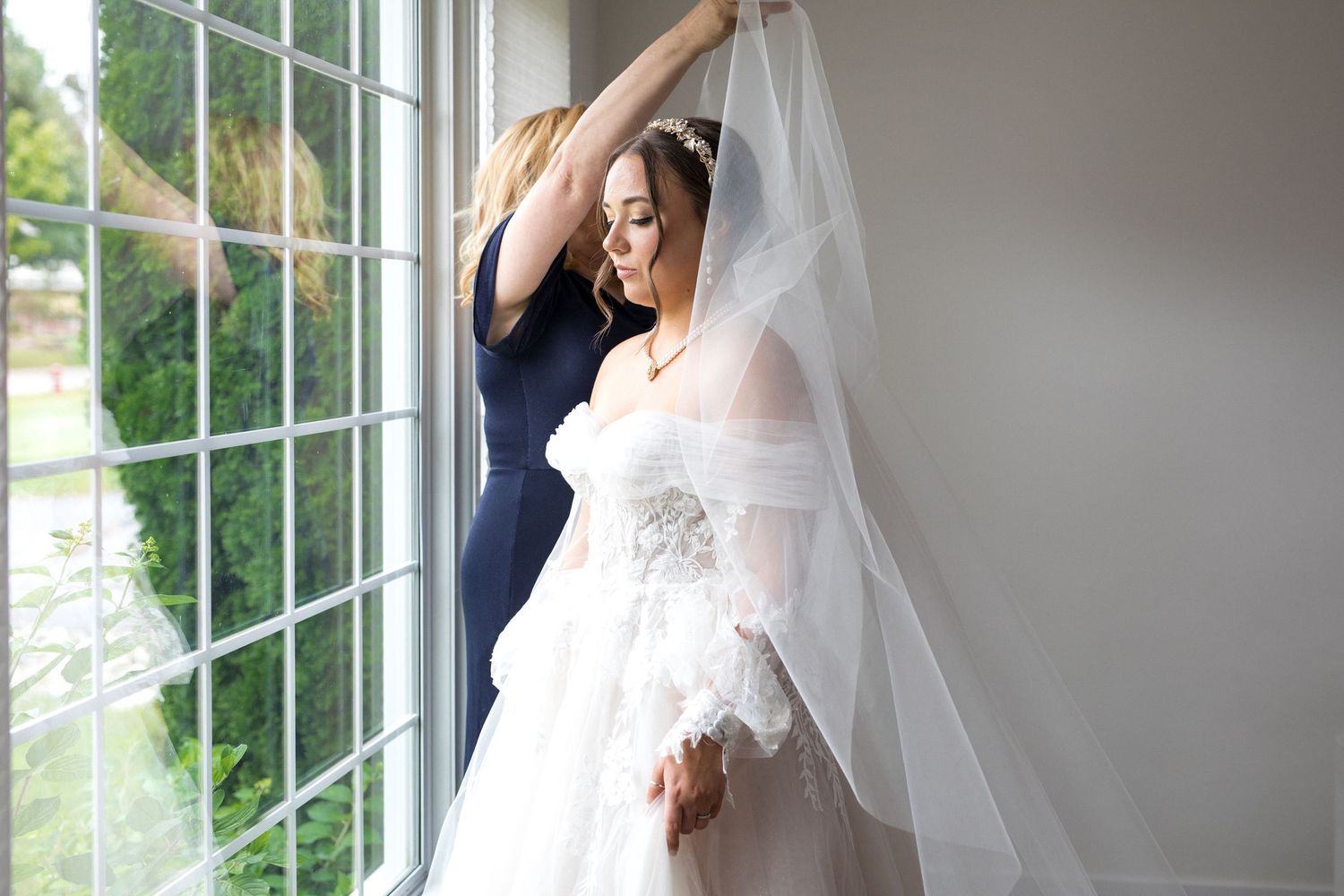 A bride in a flowing white veil poses gracefully by large windows in natural light during a wedding photoshoot.