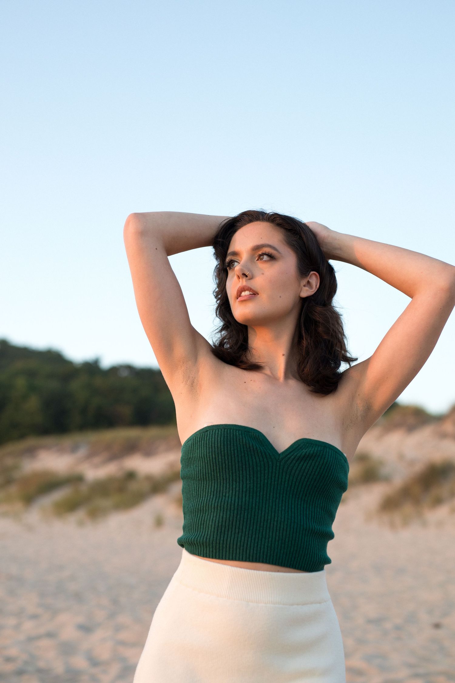 Woman in green strapless top and white skirt poses with hands behind head on sandy beach at sunset.