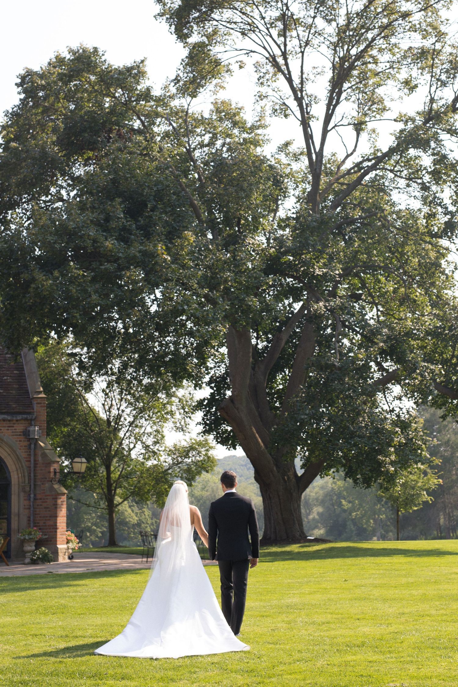 A wedding couple stands on a sprawling green lawn with mature trees and historic buildings in the background.