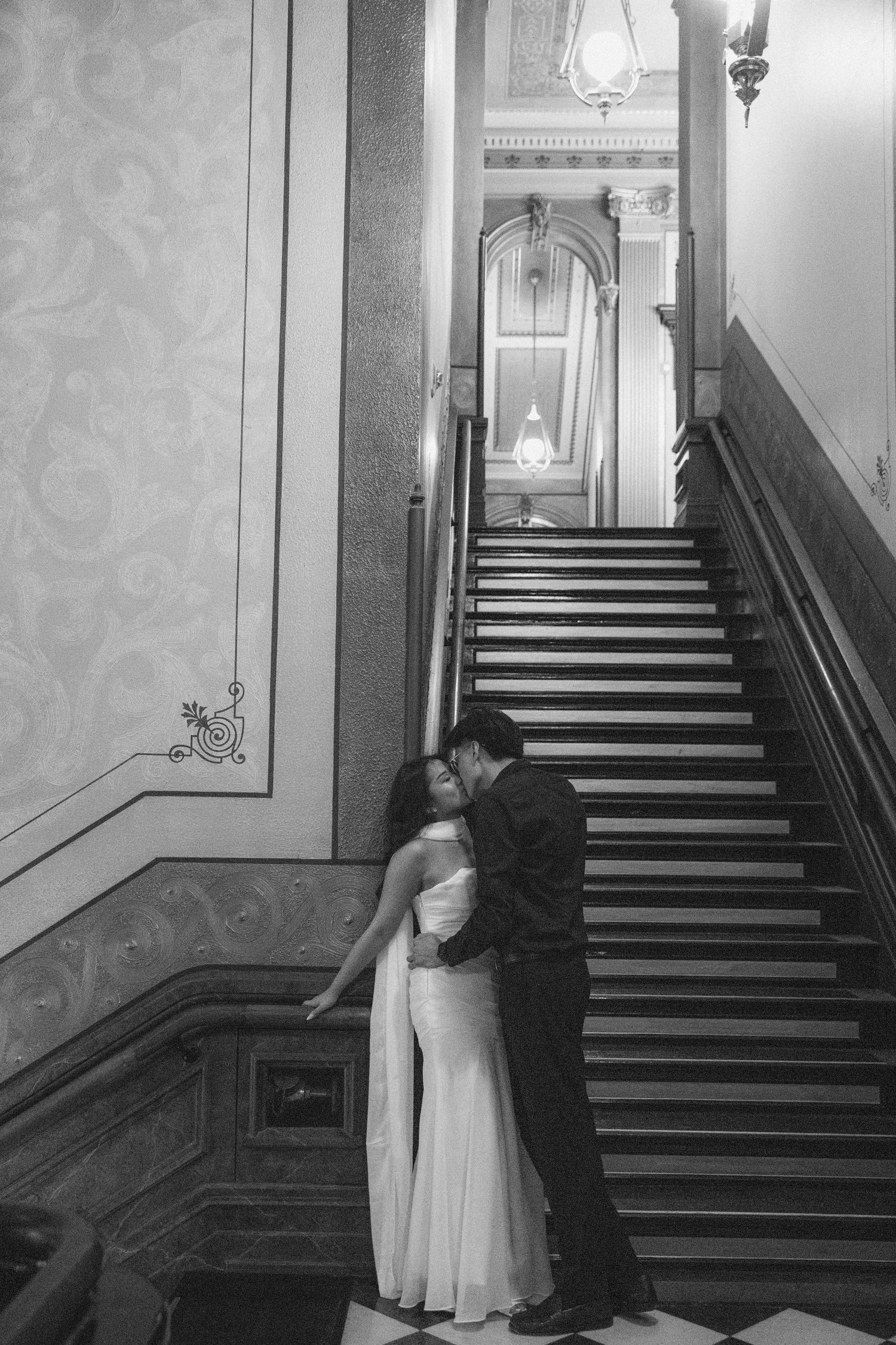 Black and white photo of a couple sharing a romantic kiss at the base of an elegant grand staircase in a formal venue.