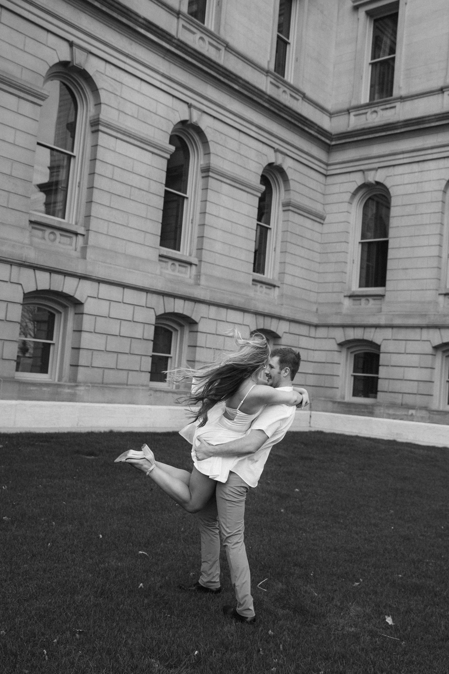 Black and white photo of a couple embracing and laughing in front of a classic stone building on a grassy lawn.