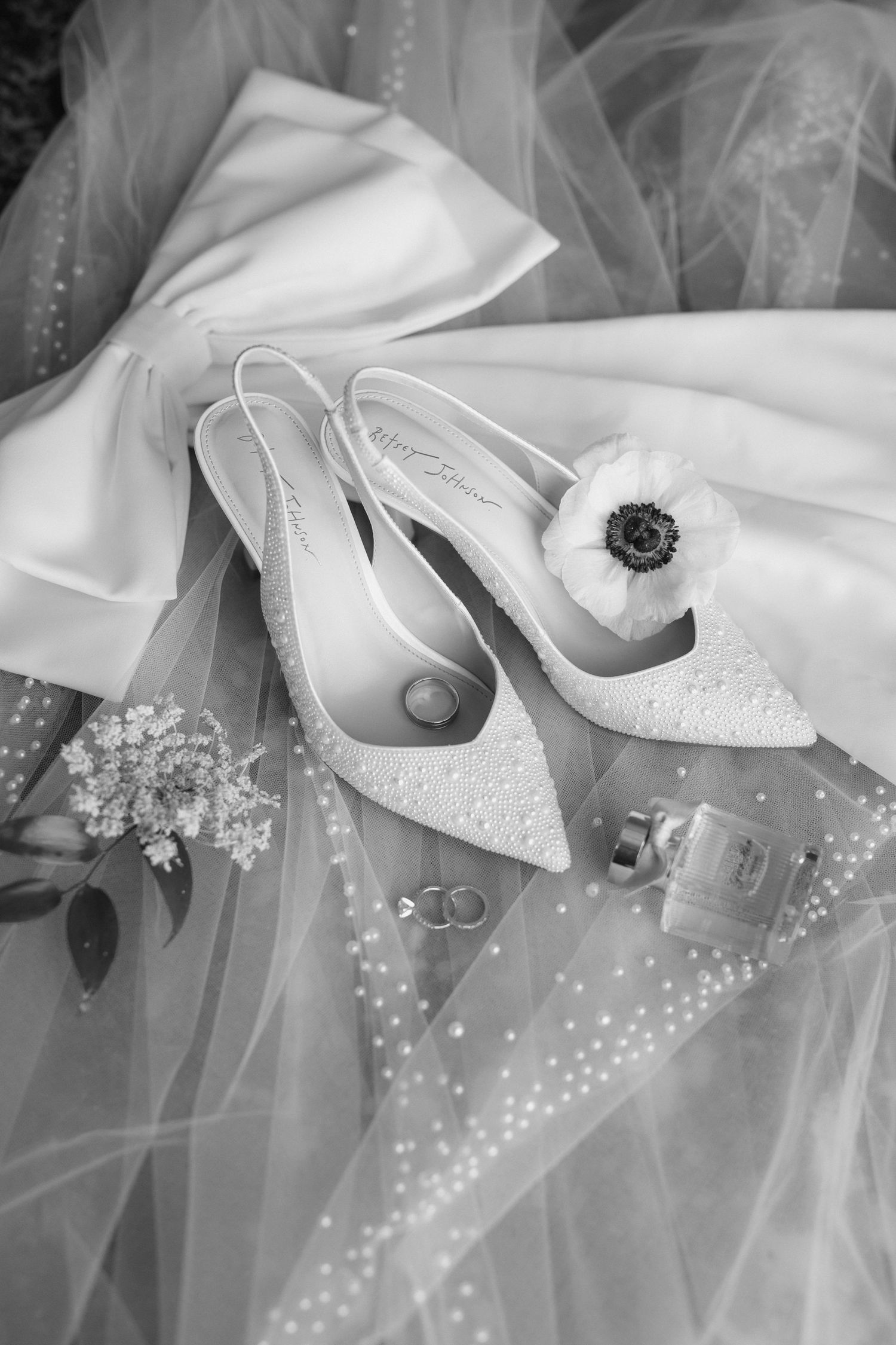 Black and white photo of bridal heels with pearl details, wedding rings, perfume, and floral bouquet on a tulle gown.