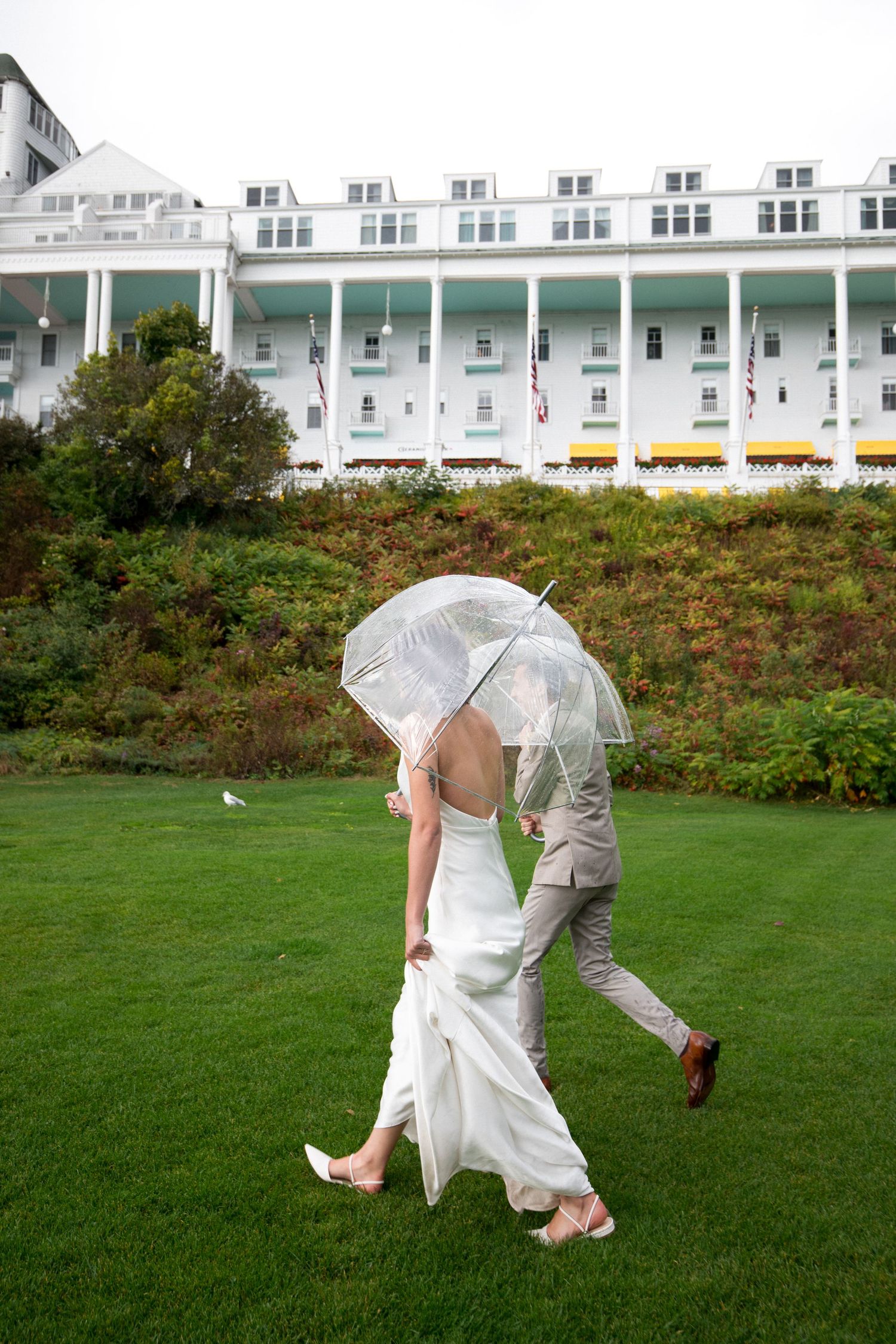 Two people in white outfits walk together under a clear umbrella on a green lawn with buildings in the background.