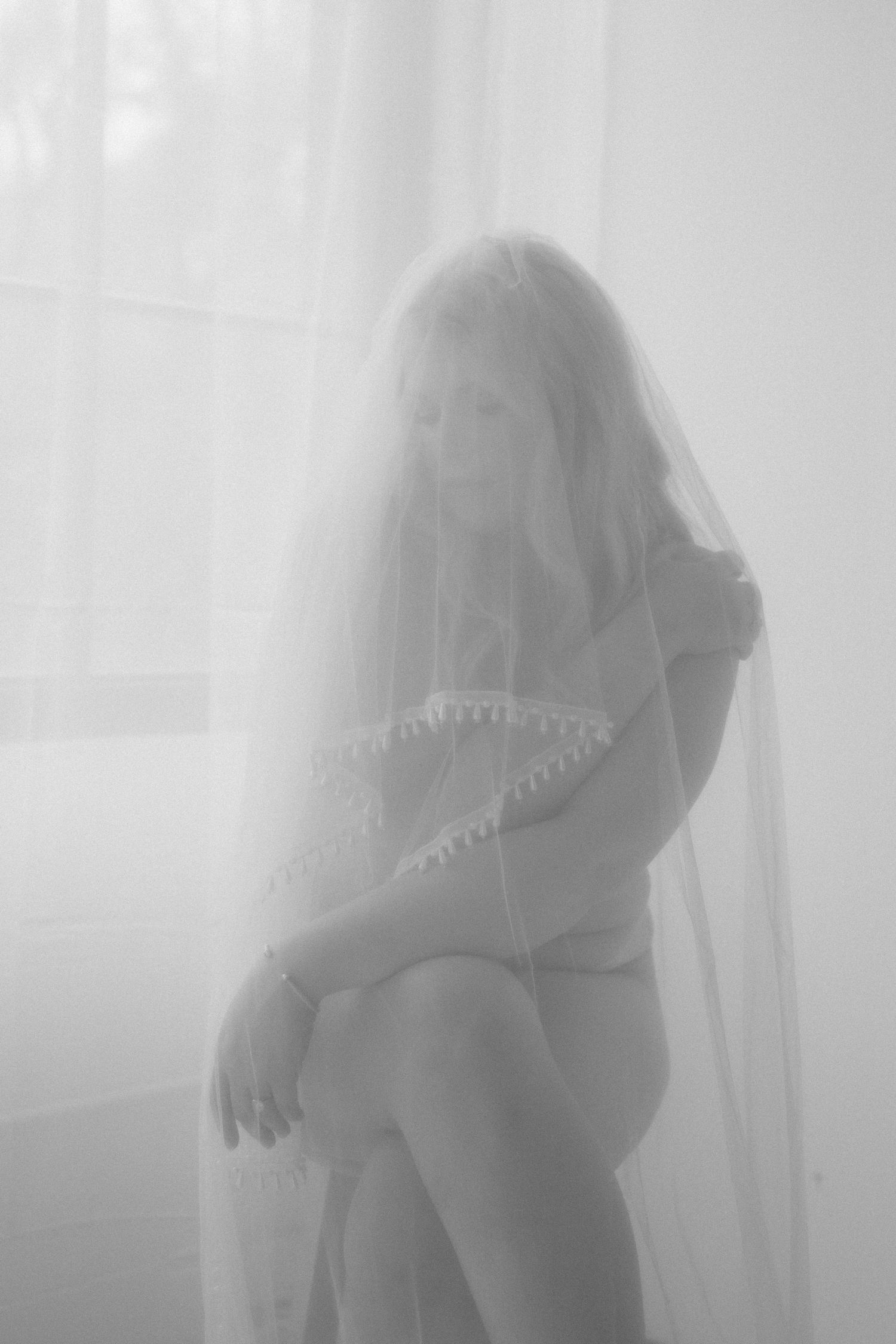 Black and white photo of a bride in a wedding veil and lace dress seated gracefully near a bright window.