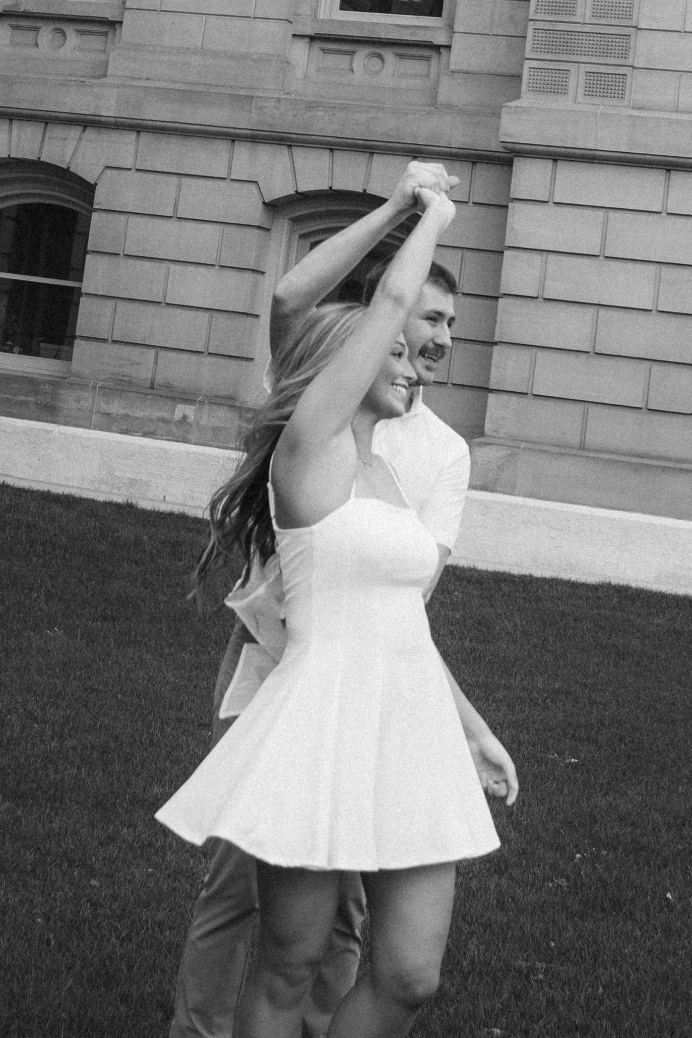 A joyful couple dancing outdoors in black and white, woman in white mini dress twirling.
